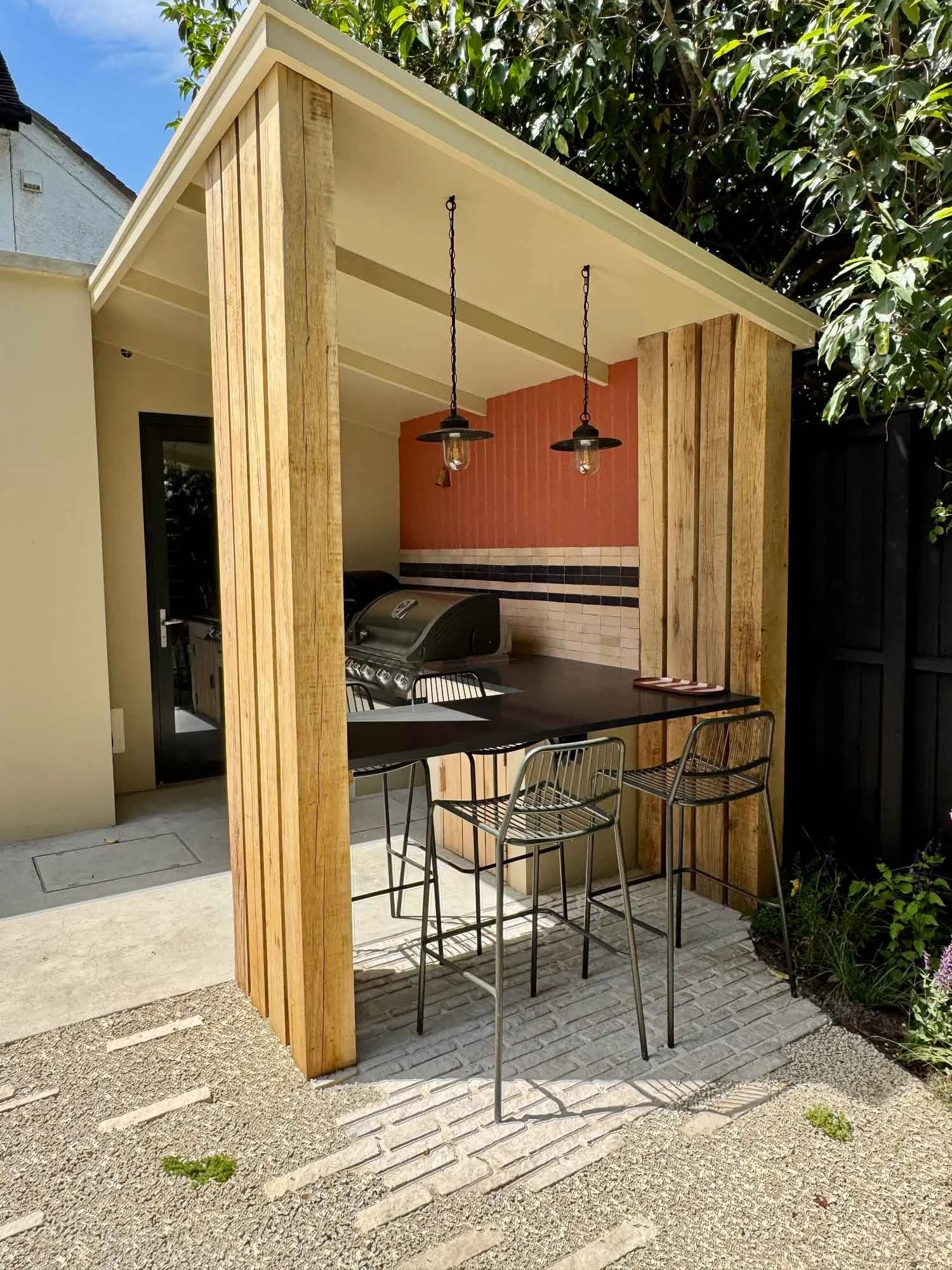 Outdoor bar area with a black countertop, two metal bar stools, hanging pendant lights, and a built-in grill, surrounded by wooden panels and greenery.