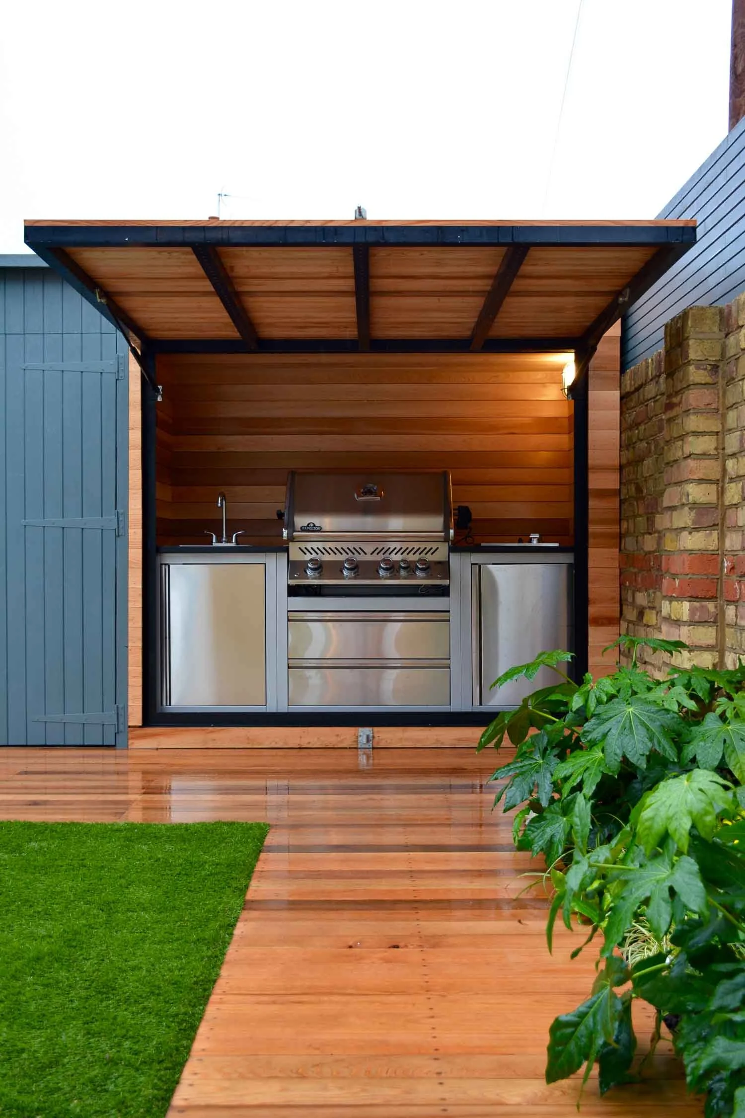 A backyard outdoor cooking station with a stainless steel grill, sink, and storage cabinets within a wooden shed with an open front, surrounded by a wooden deck, green grass, and plants.