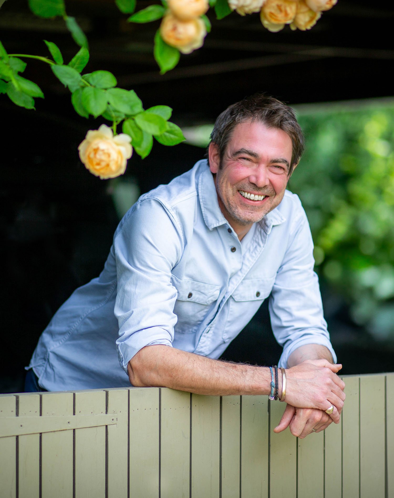 Smiling man with short dark hair leaning on a wooden fence under yellow climbing roses in a bright outdoor setting