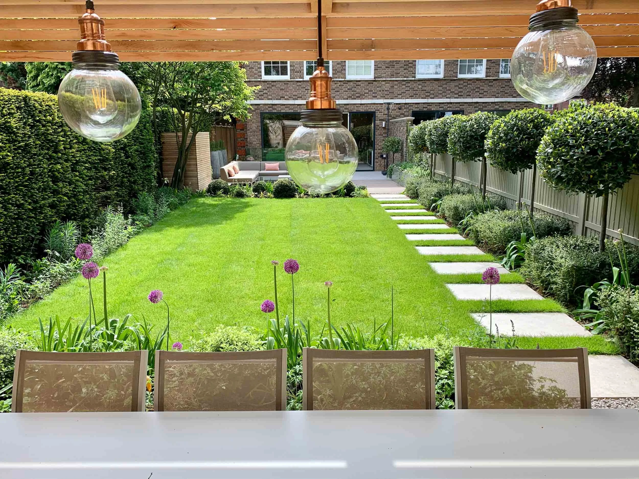 A well-maintained backyard garden viewed from a patio or deck, with a white table and four chairs in the foreground, a green lawn with purple flowers, stepping stones on the right side, and a sitting area with outdoor furniture near a brick house in 