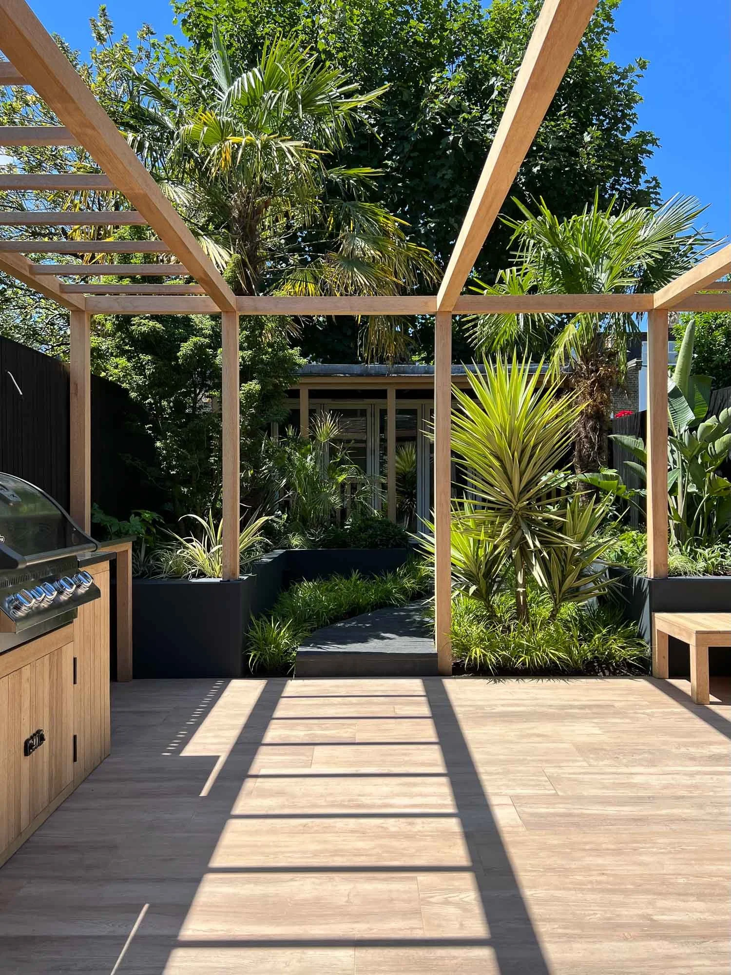 An outdoor patio area under construction with a wooden frame structure, surrounded by lush green tropical plants and trees, with shadows cast by the wooden beams on a light-colored wooden deck.