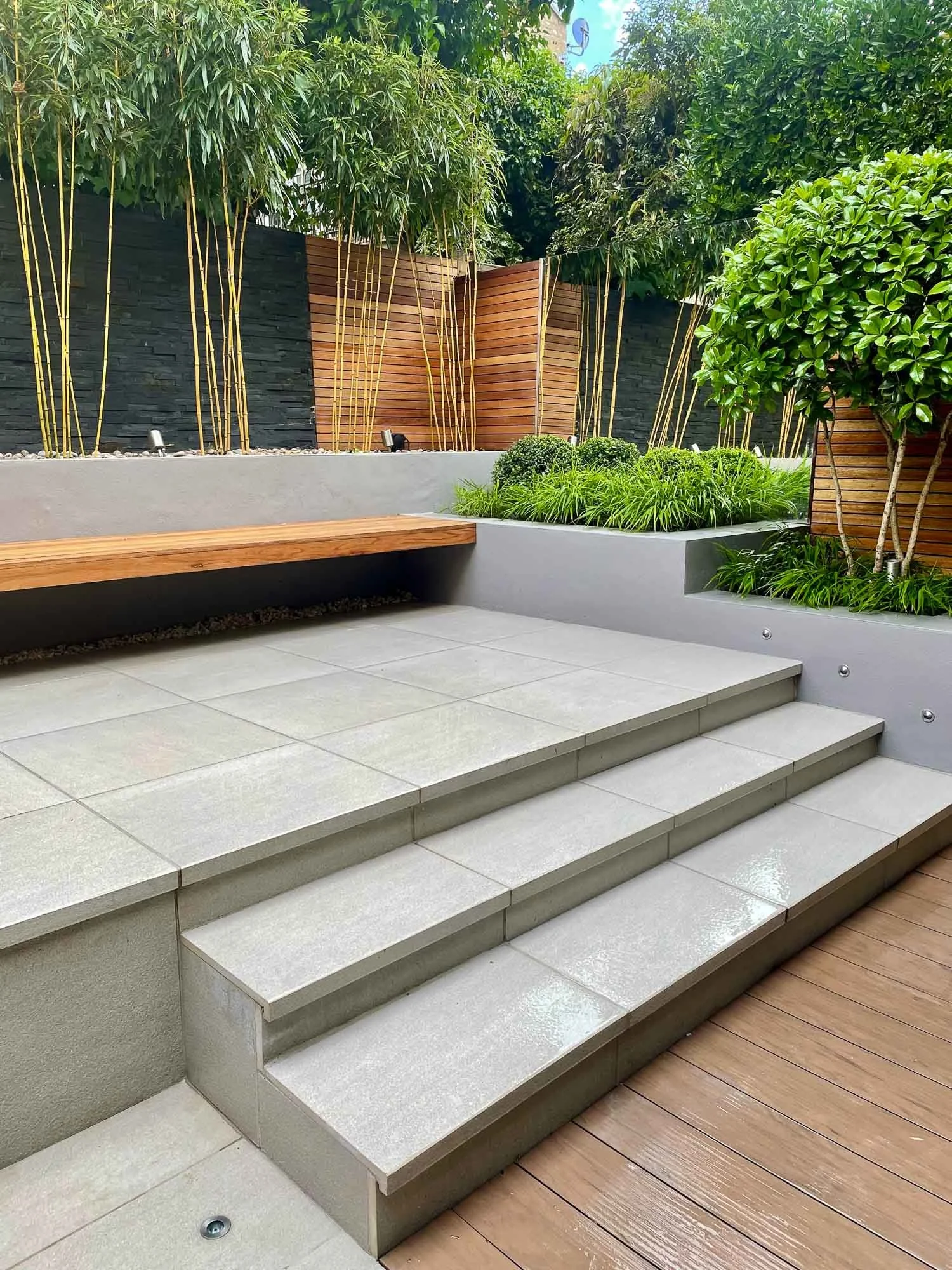 Modern outdoor patio with tiled steps, wooden deck, and lush green plants enclosed by black, brown, and gray wooden fences.
