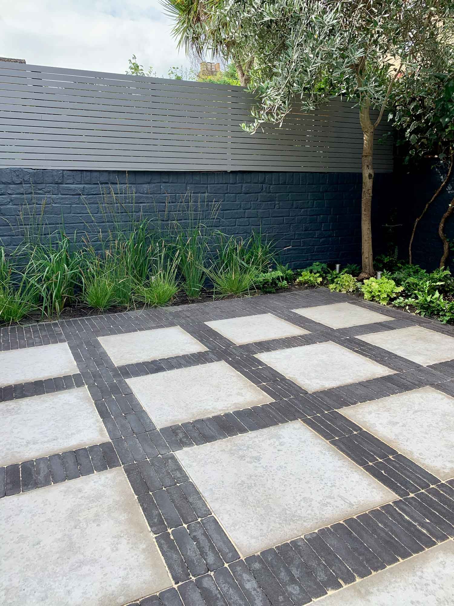 Modern outdoor patio with large square concrete tiles surrounded by dark bricks, a wooden fence, trees, and greenery.