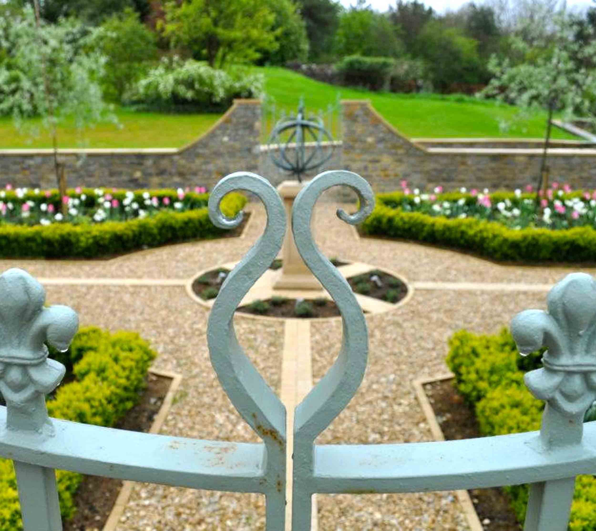 View of a garden with manicured flower beds and a stone wall in the background, captured through a decorative white metal gate with a curled design.