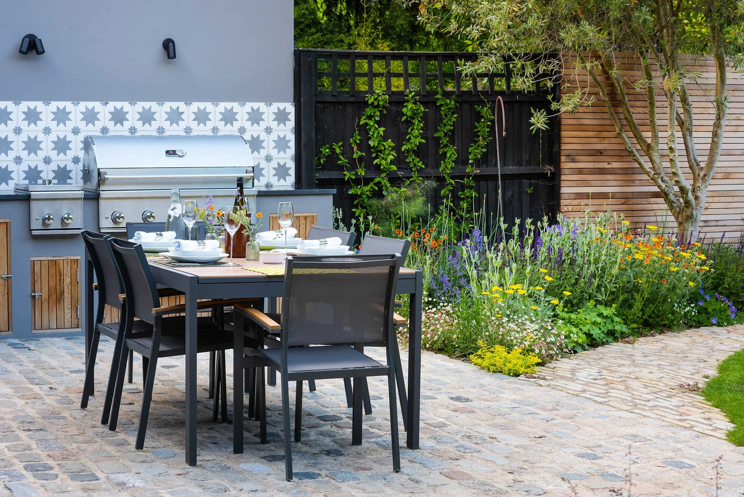 Outdoor dining set on a cobbled patio next to an outdoor kitchen and a colourful garden border with purple salvia