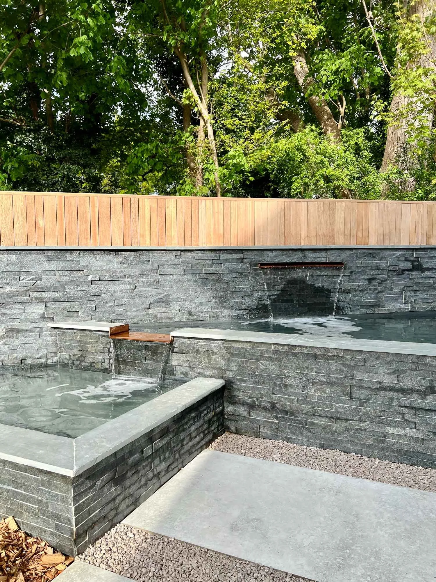 A modern outdoor water feature with black brick walls, a water spout, and a small pool on a concrete patio, surrounded by lush green trees.