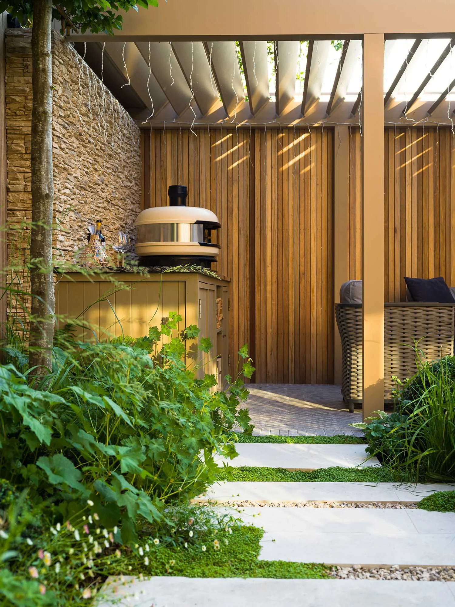 Close-up of garden path with stone stepping stones and ground cover leading towards an outdoor kitchen and pizza oven