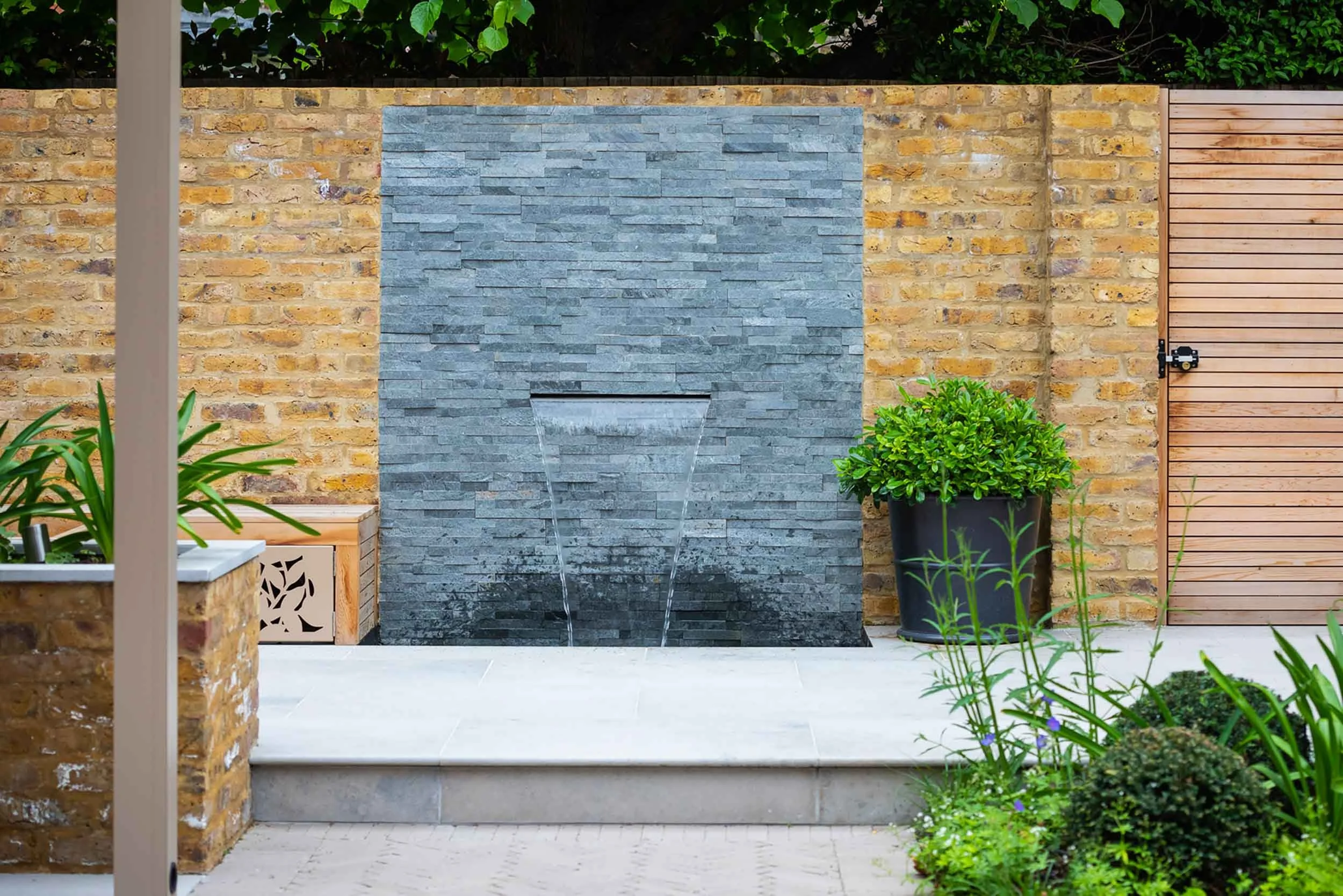 Contemporary slate blade water feature falling into a pool on a stone patio with yellow brick walls and lush planting