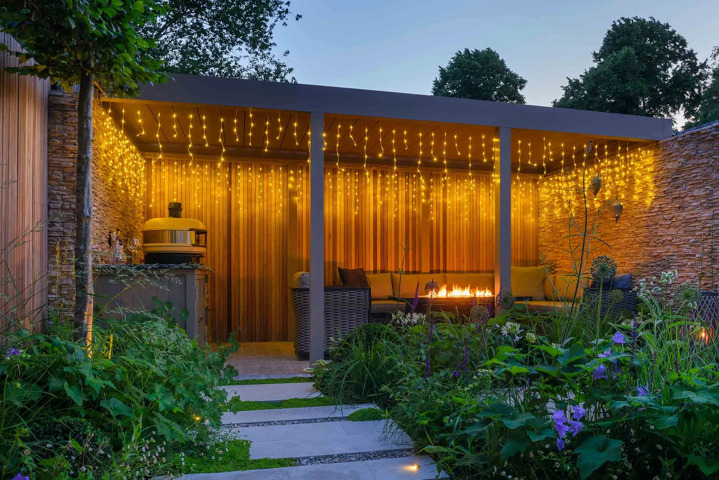 Evening view of the garden pergola and seating area, illuminated by warm yellow string lights and a flickering, linear outdoor fire