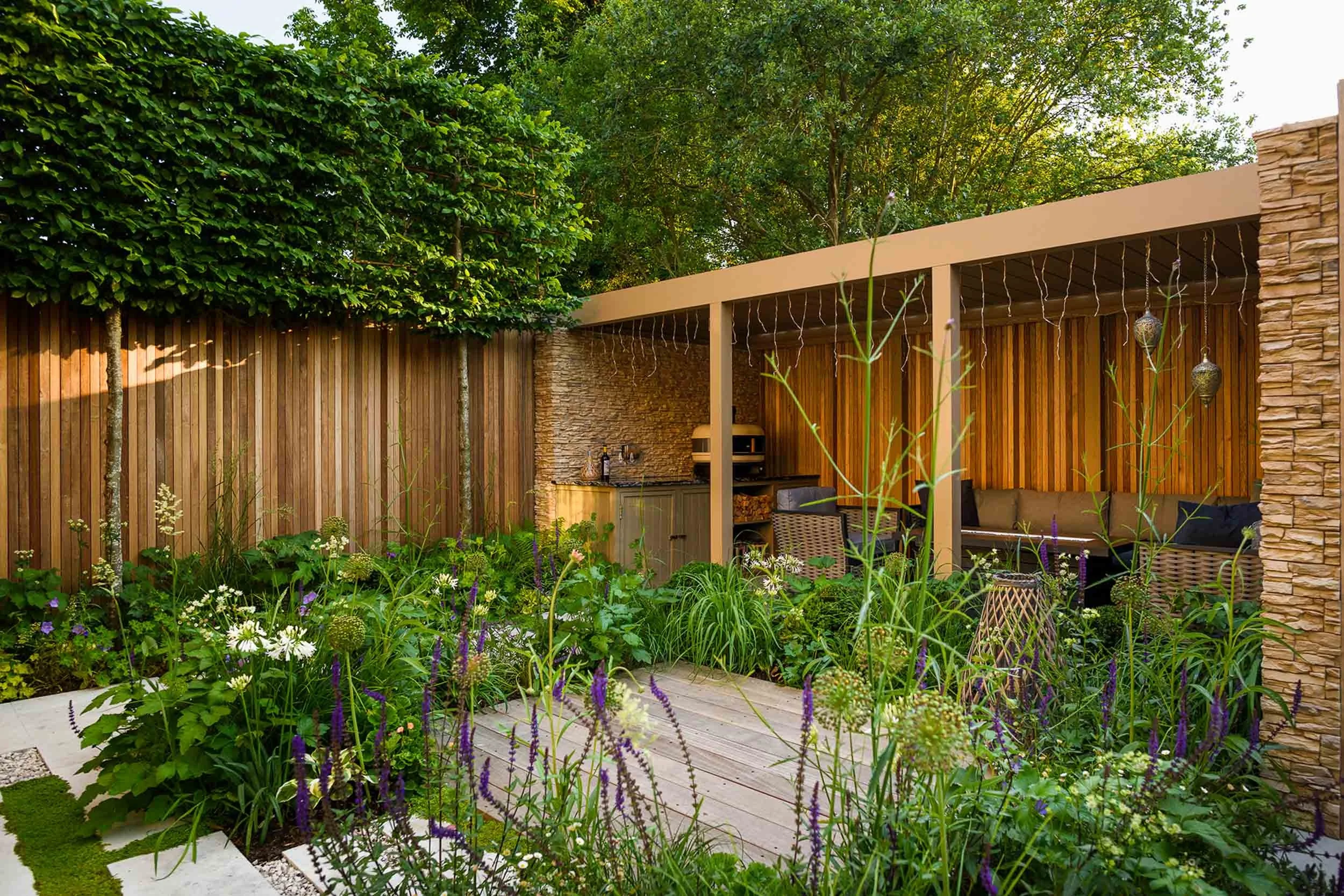 Wide garden view featuring a pergola, vertical timber fence, high leafy hedge, an outdoor kitchen, and densely planted flowerbeds