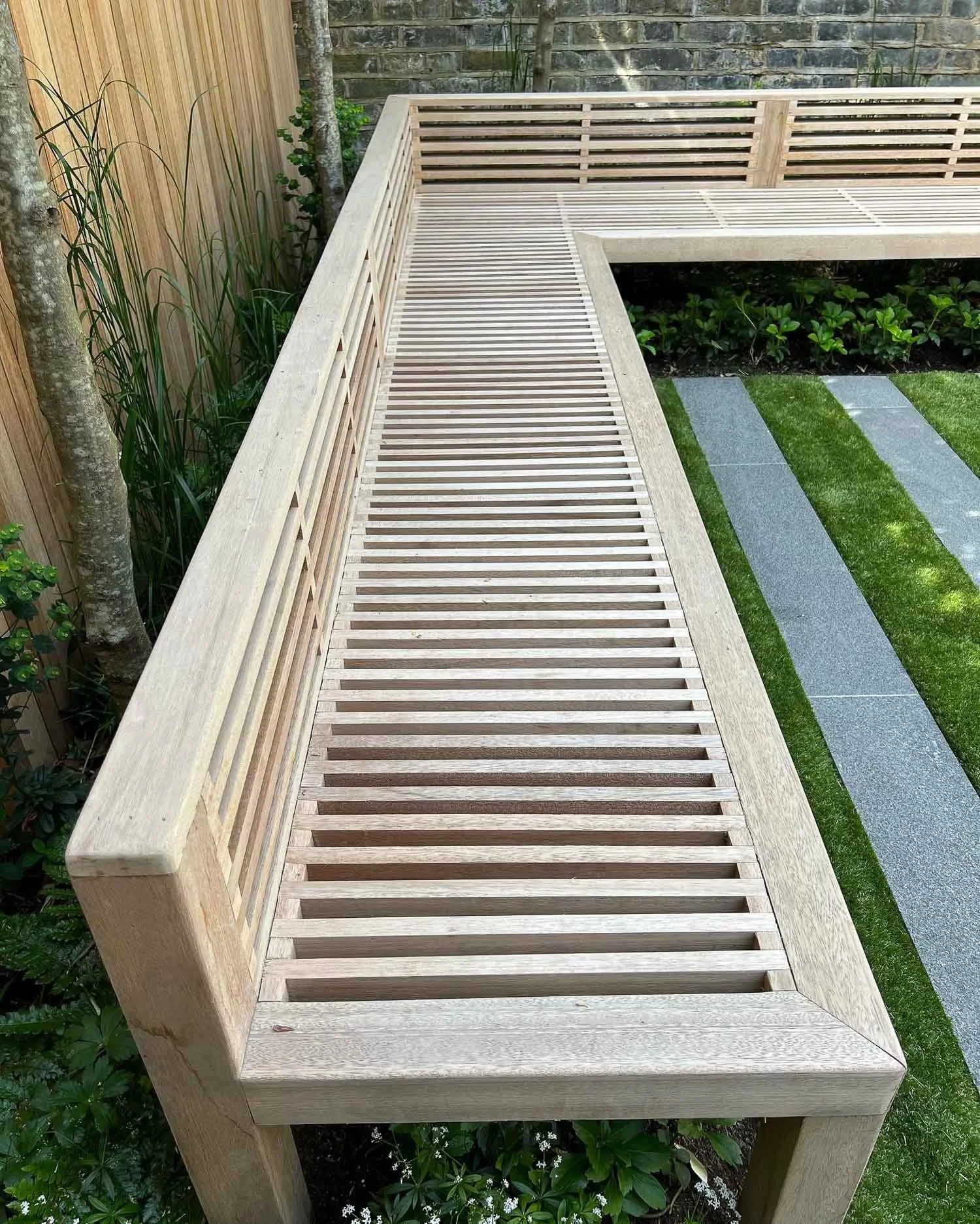 Wooden outdoor bench with slatted top and side railing, situated in a garden with greenery and stone pathway.
