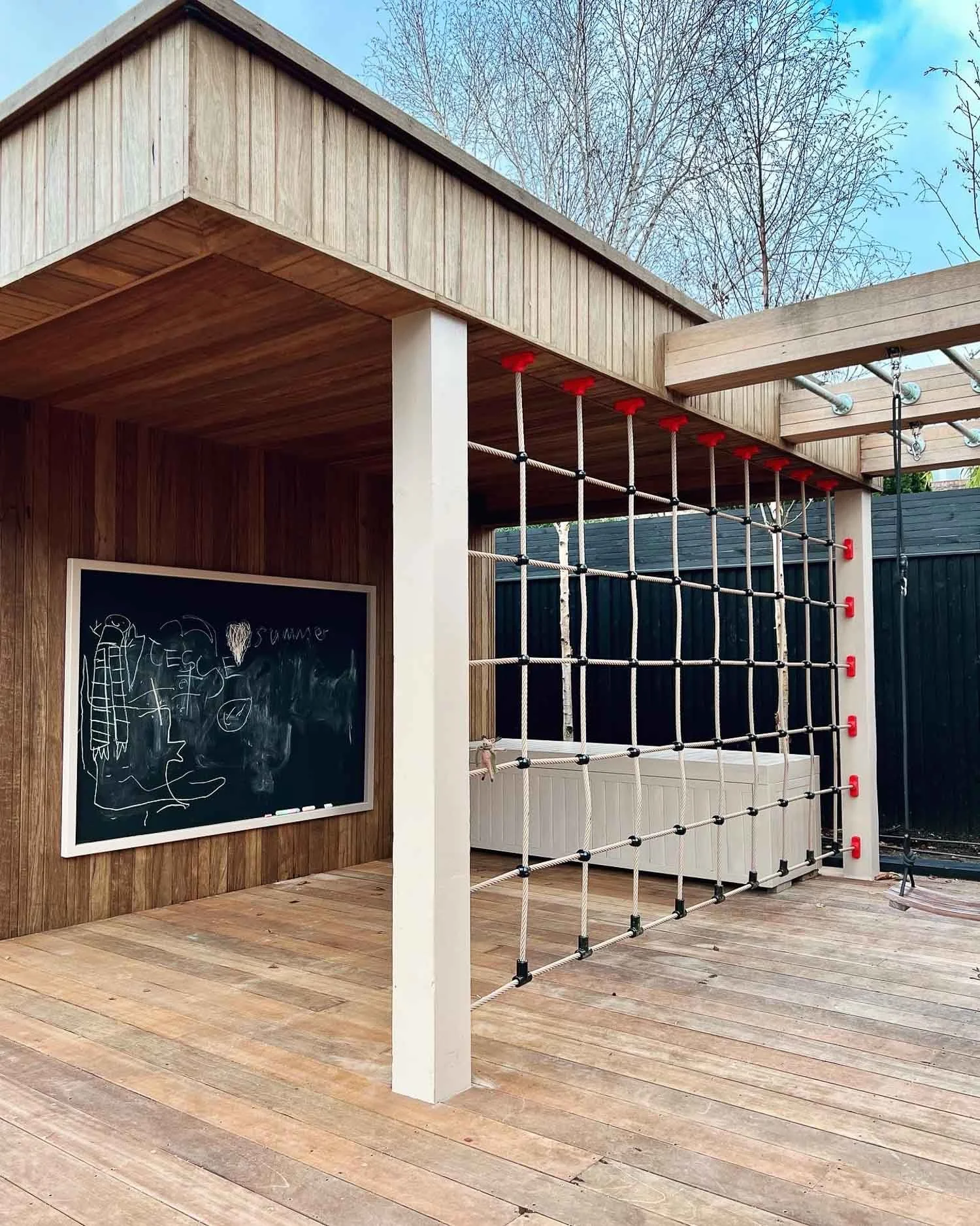 Wooden outdoor patio with climbing rope structure, black chalkboard with drawings, and a hot tub. Trees are visible in the background.