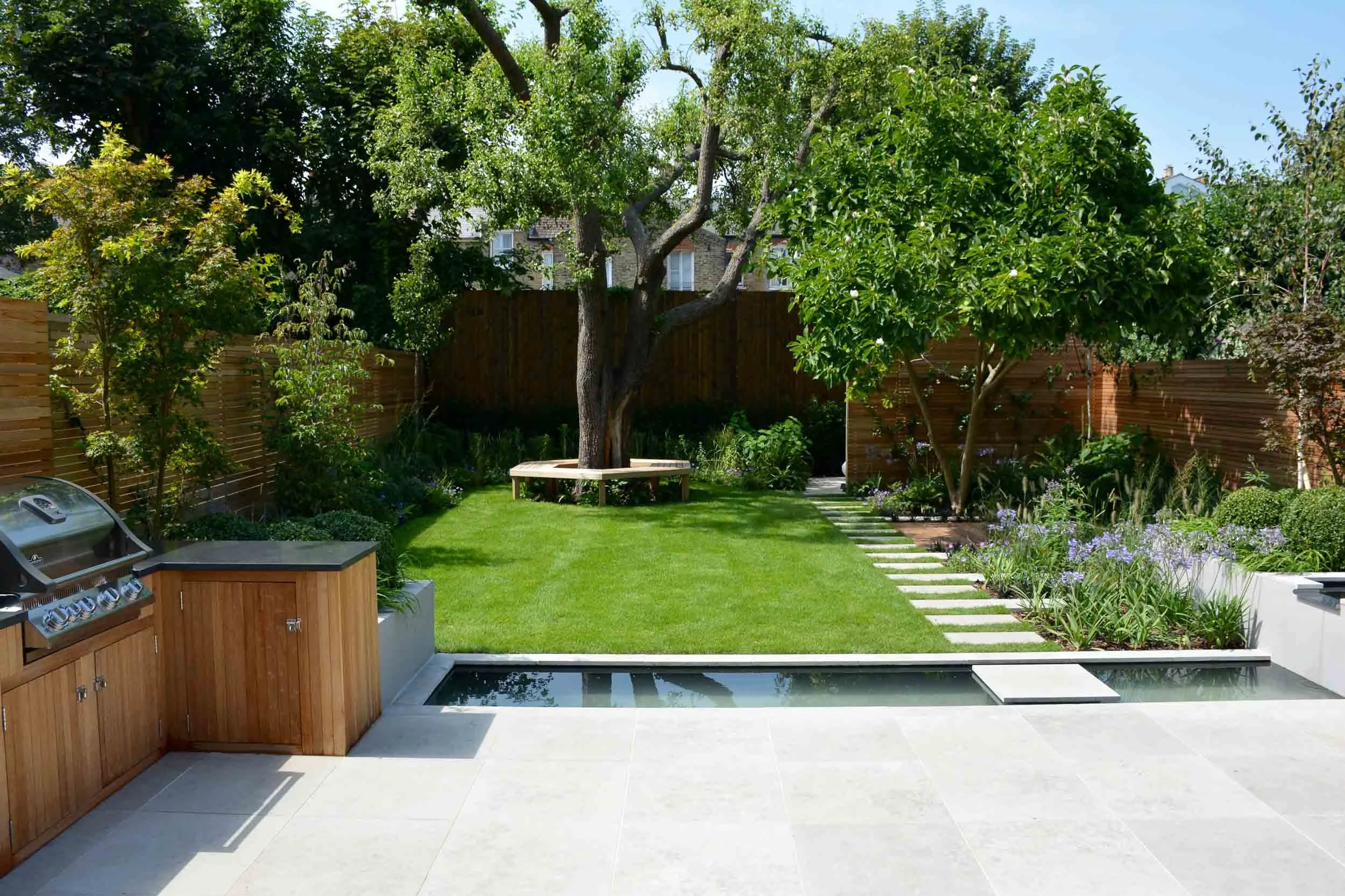 A backyard garden with a grassy lawn, trees, a wooden bench, and a stone pathway, bordered by flower beds and a wooden fence, with a built-in outdoor kitchen area in the foreground.