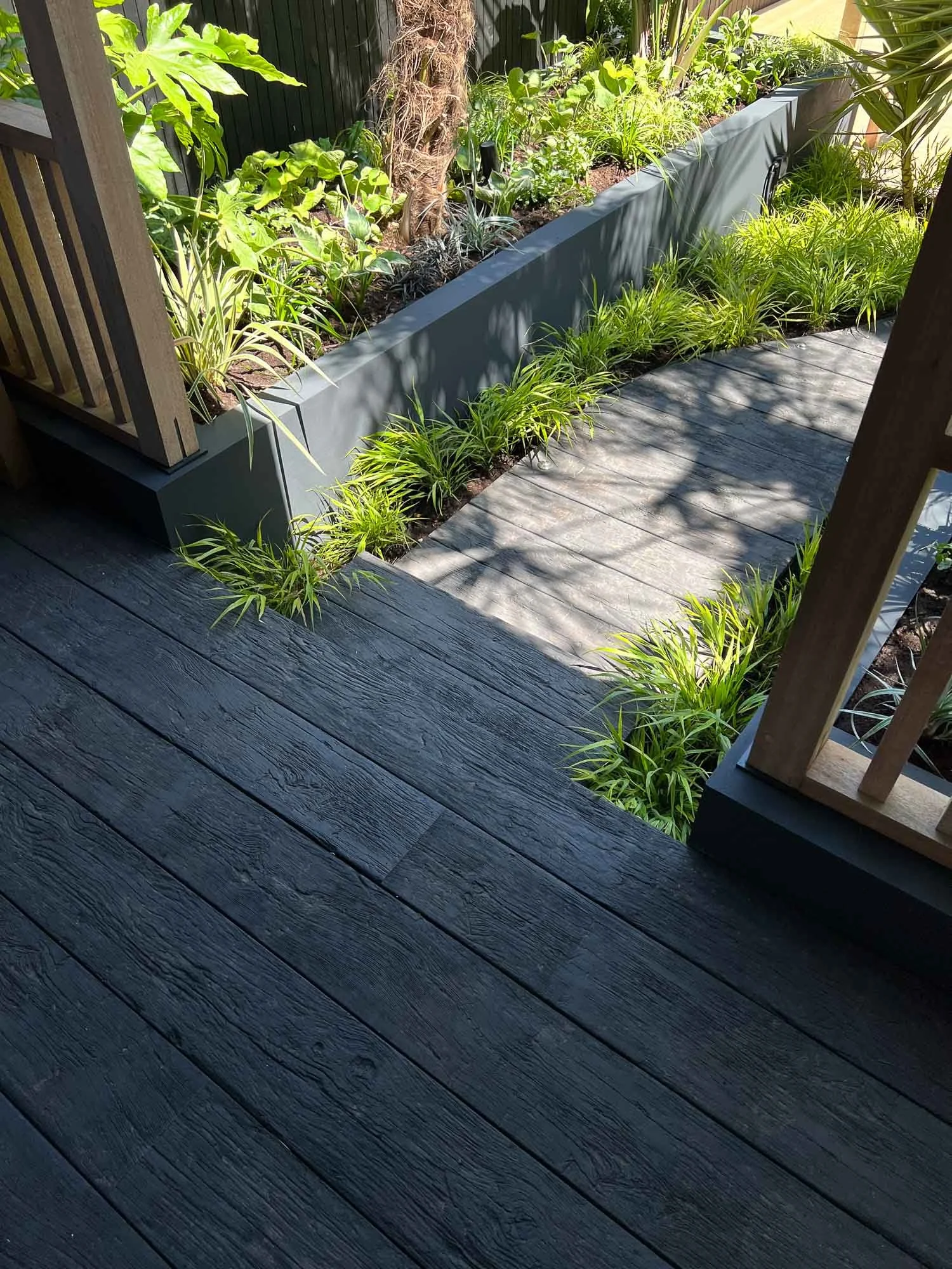 A backyard with a wooden deck and a raised garden bed filled with green plants, with shadows cast by nearby trees.