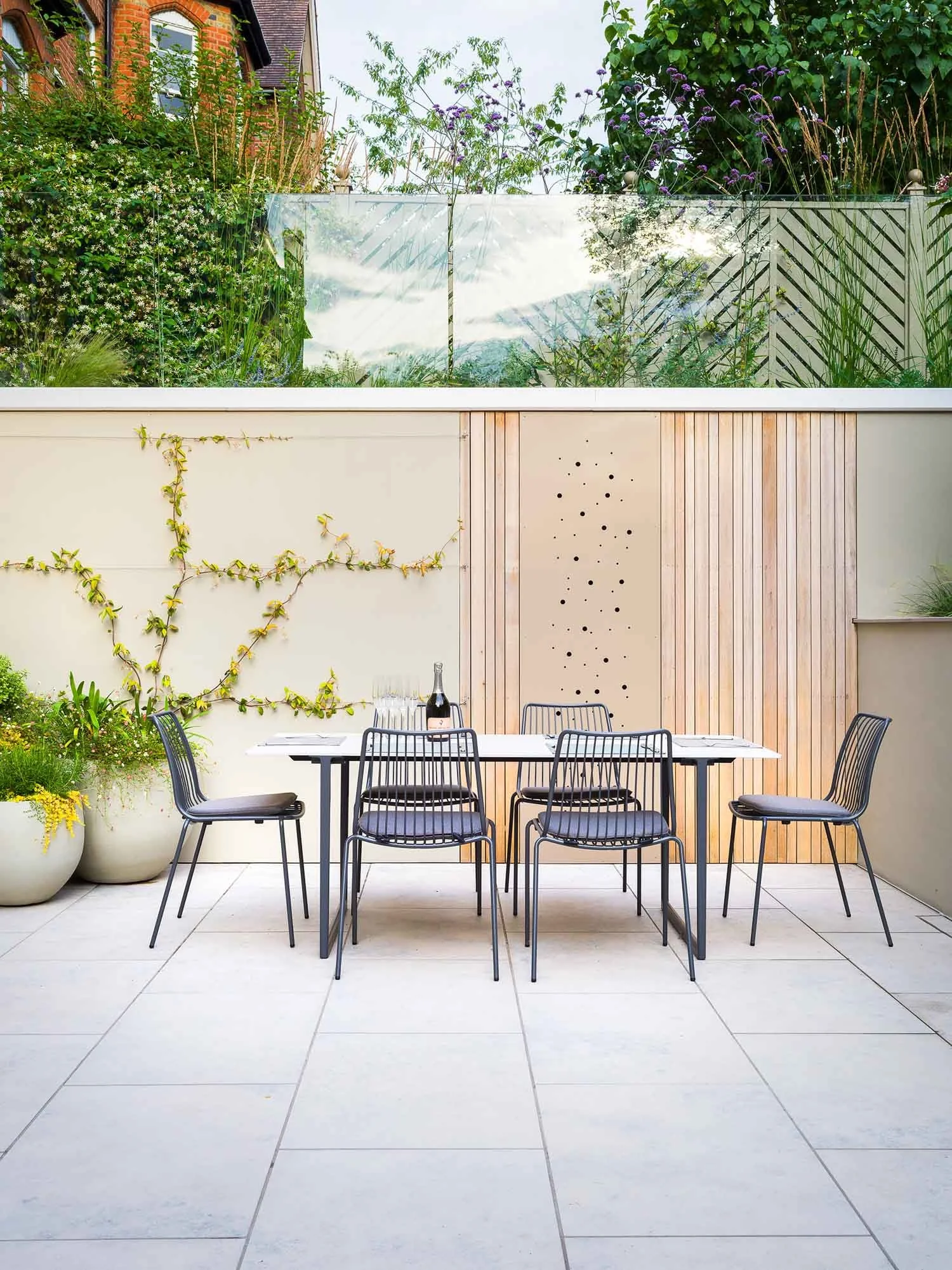 Dining area on a stone terrace featuring a modern black metal table and chairs surrounded by raised rendered planters and green foliage