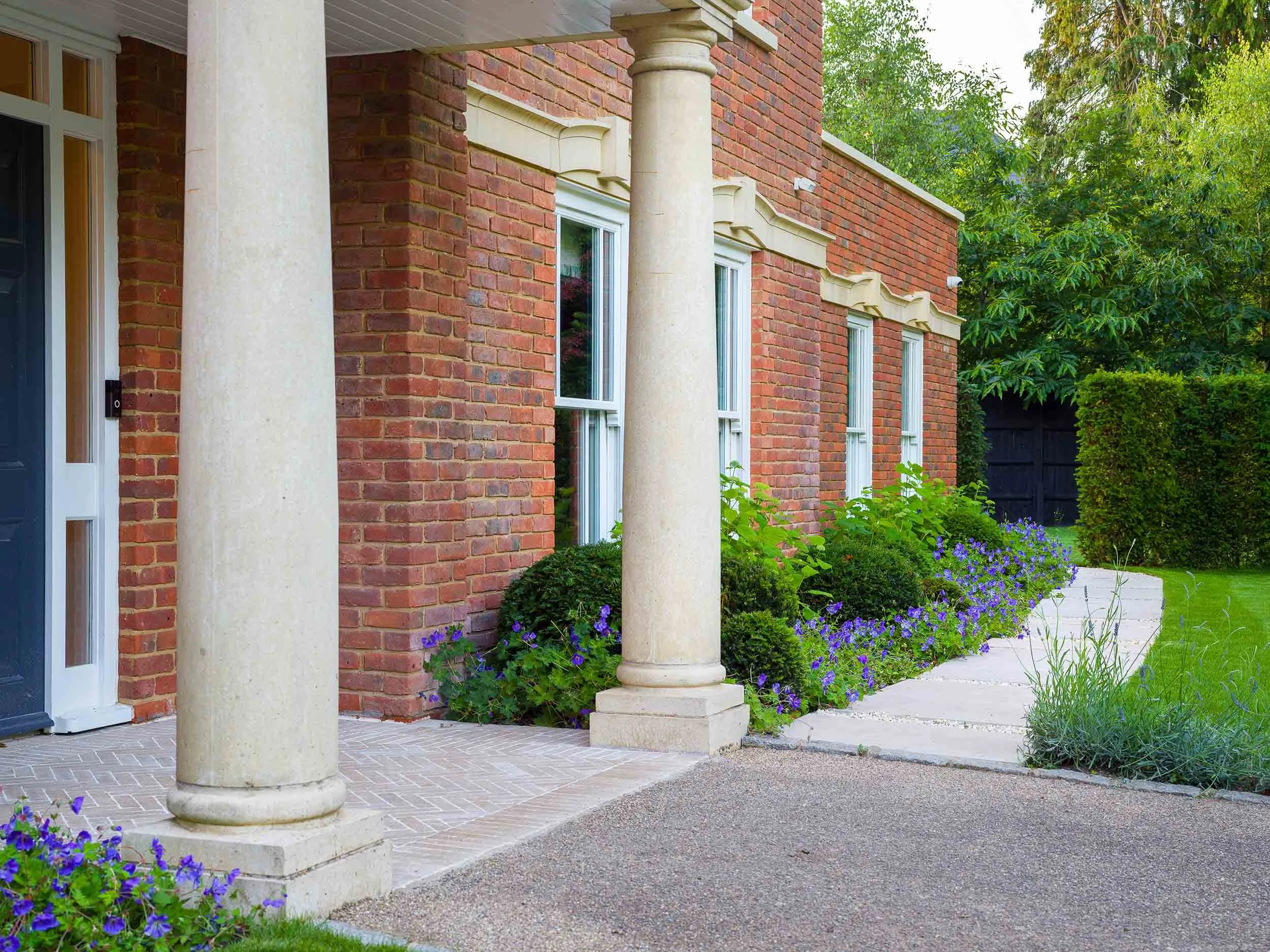 Stone pillars and red brick house with white windows overlooking a stone path lined with blue geranium flowers