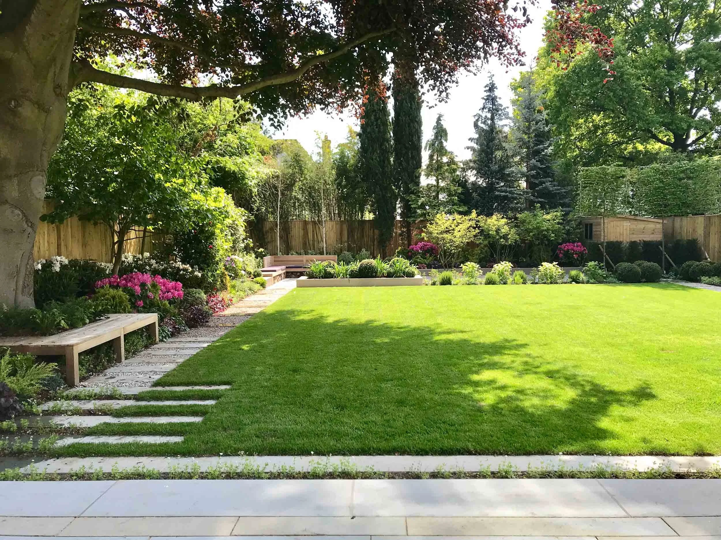 A well-maintained backyard garden with a lush green lawn, a variety of trees and flowering plants, a curved pathway with stepping stones, and a wooden bench along the walkway. Tall trees and a wooden fence surround the garden.