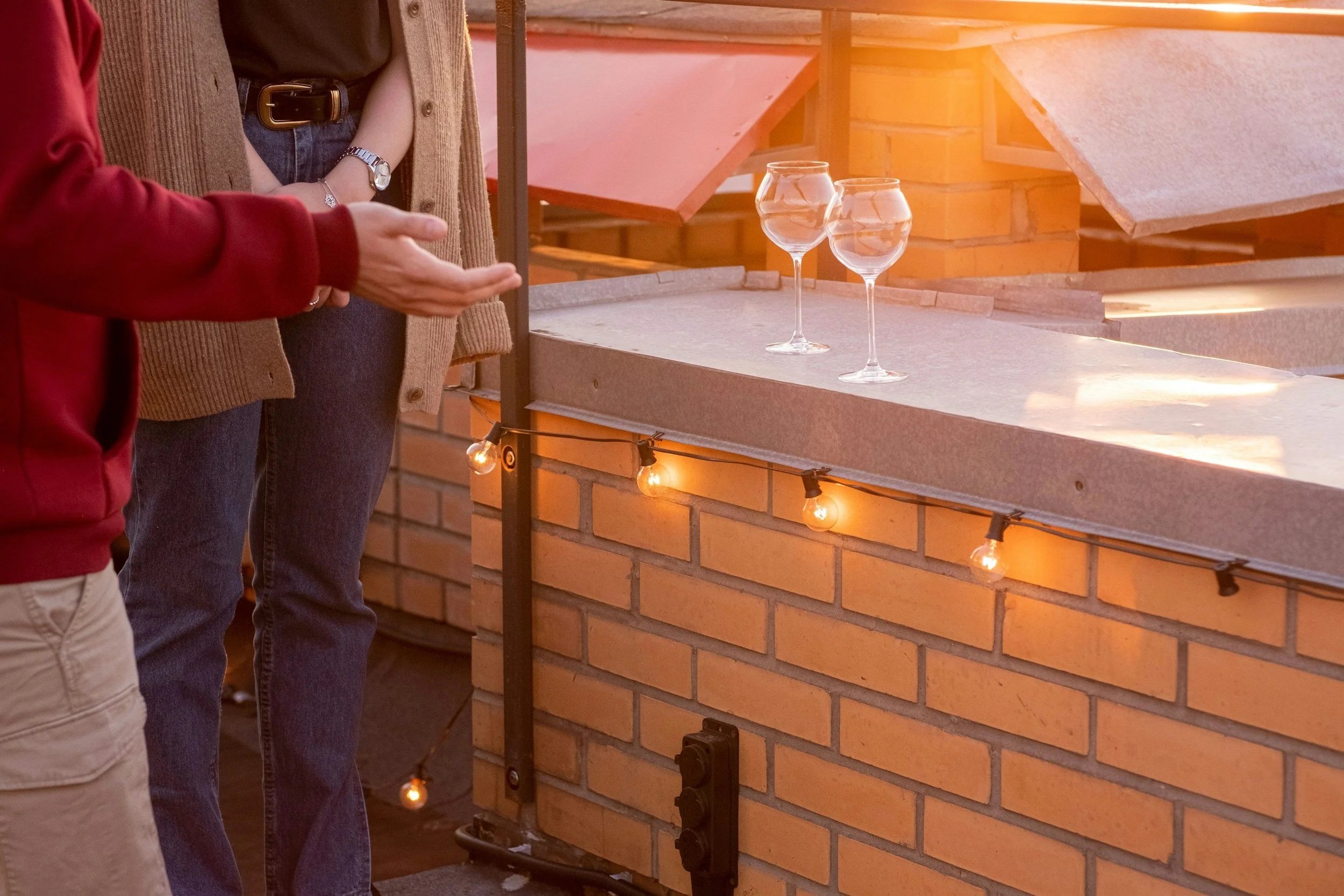 Two people standing near a specialist brick counter in Glasgow with string lights and two empty wine glasses on top, on a rooftop with sunlight.
