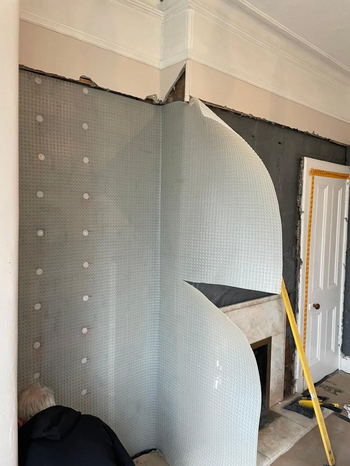 View of a partially demolished interior wall with drywall and insulation removed, exposing the structure inside a room with a fireplace and a closed door, construction tools present.
