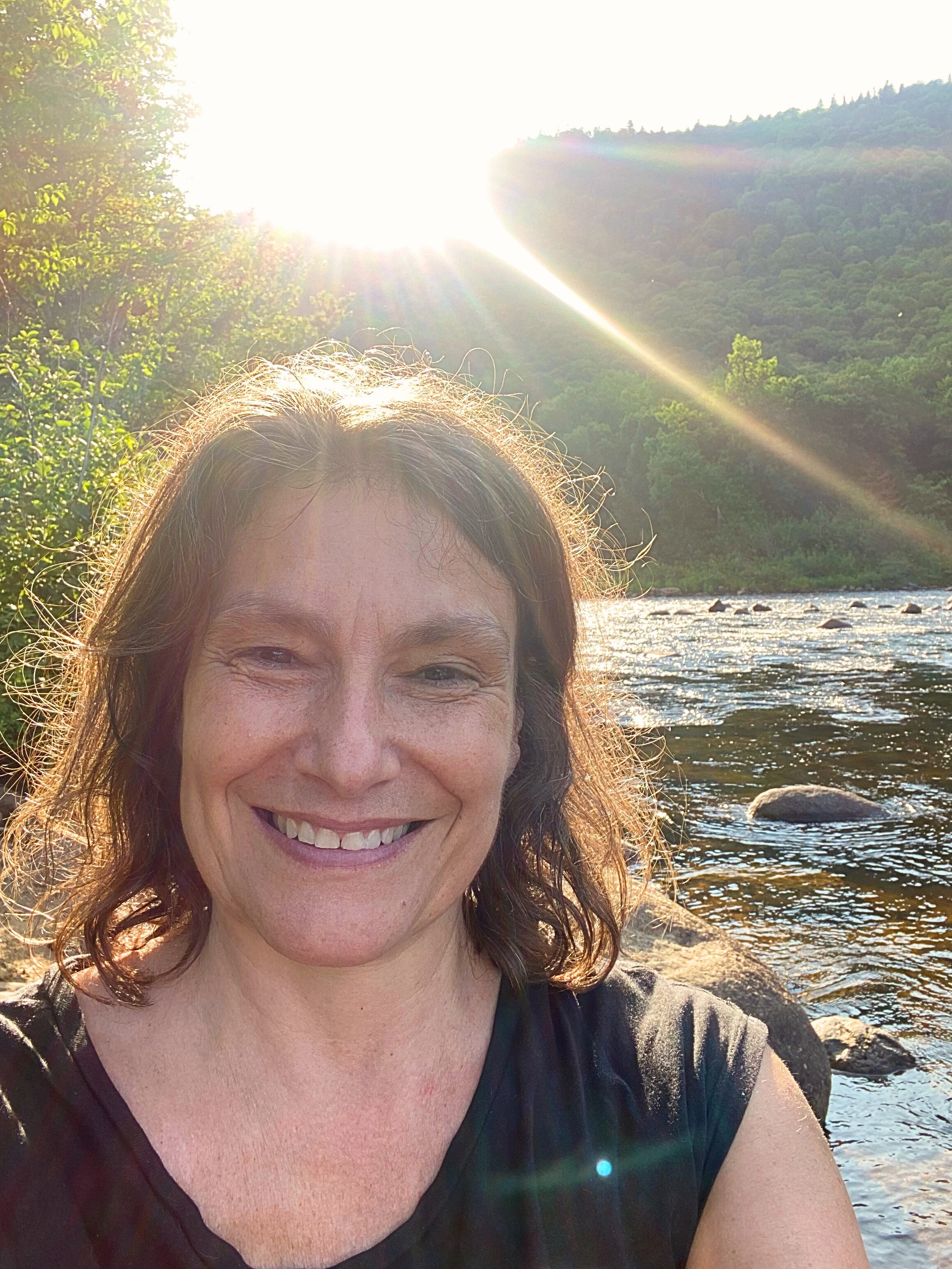 Une femme souriante près d'une rivière, avec un soleil brillant au-dessus d'une montagne verte en arrière-plan.