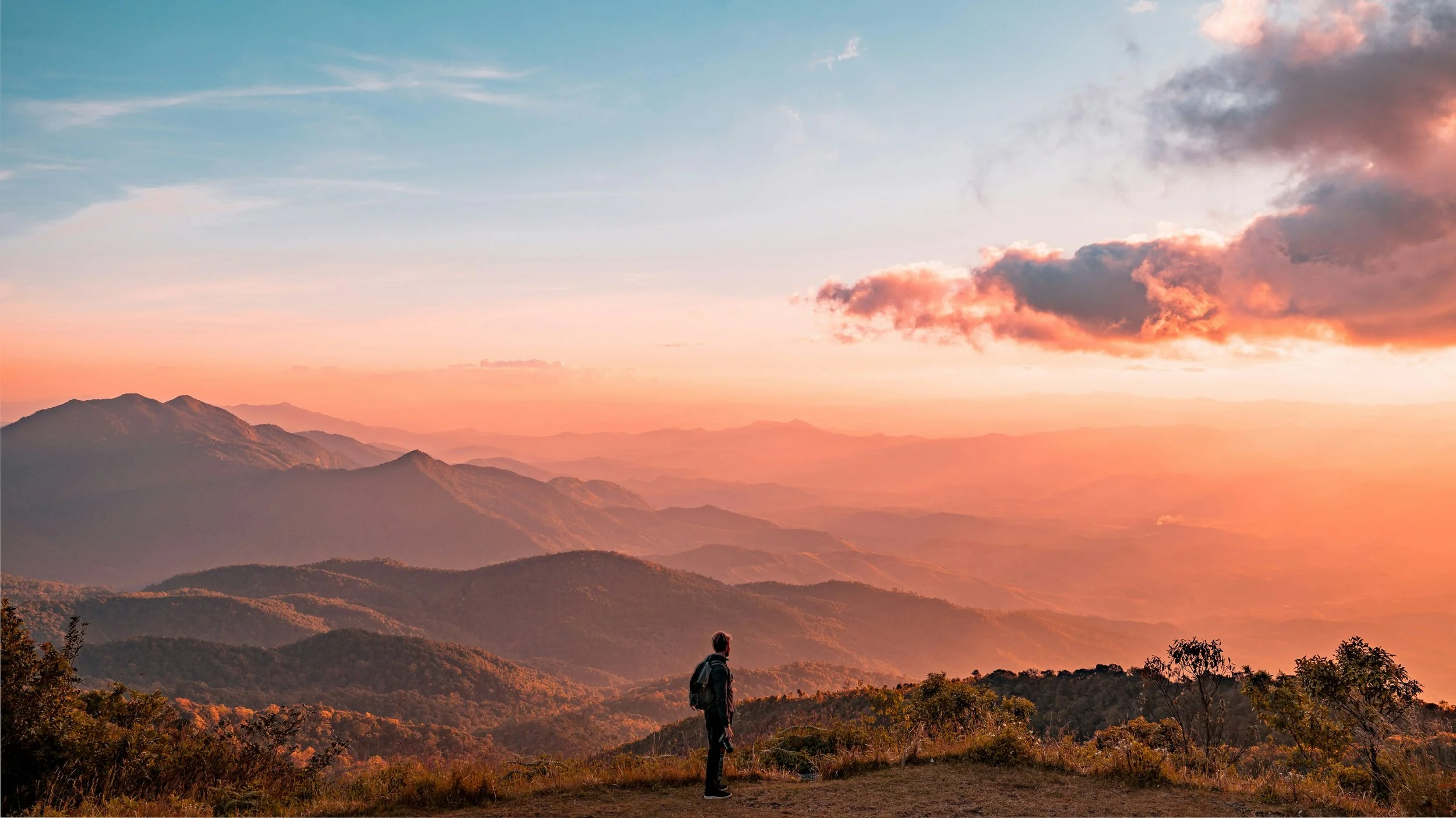 Homme regardant les montagnes lors d'un coucher de soleil avec des nuages roses et une vue panoramique.