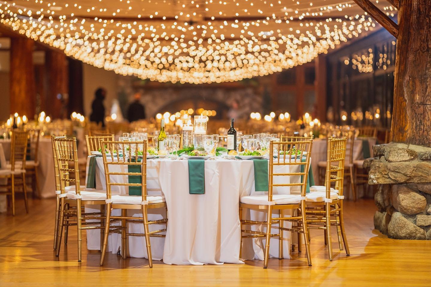 Elegant banquet hall decorated for a celebration with round tables covered in white tablecloths, gold chiavari chairs, green table runners, and table settings including wine bottles, glasses, and floral arrangements. The ceiling is adorned with hanging string lights creating a warm, festive ambiance.