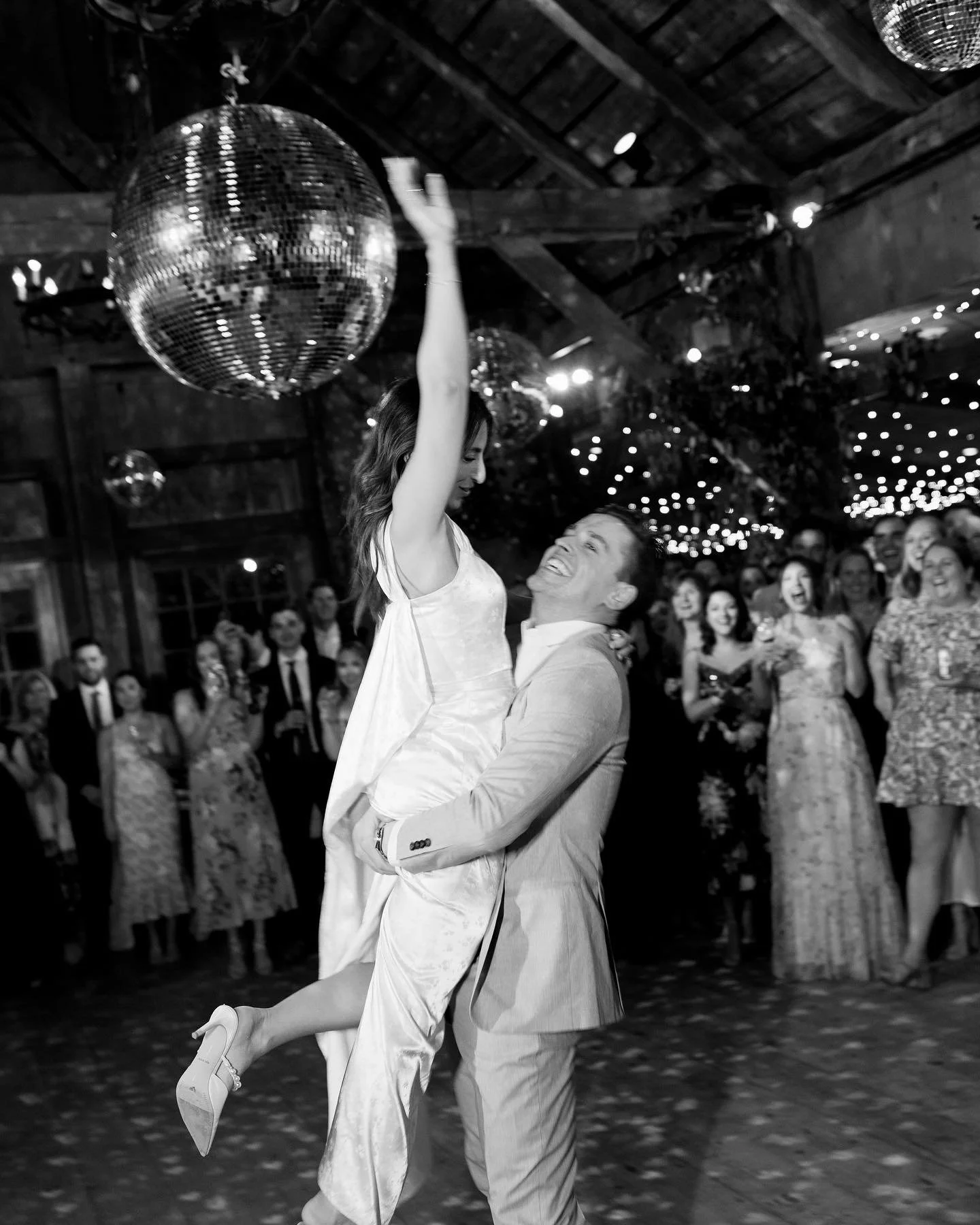 Black and white photo of a man lifting a woman during a dance at a celebration, surrounded by guests and decorated with string lights.