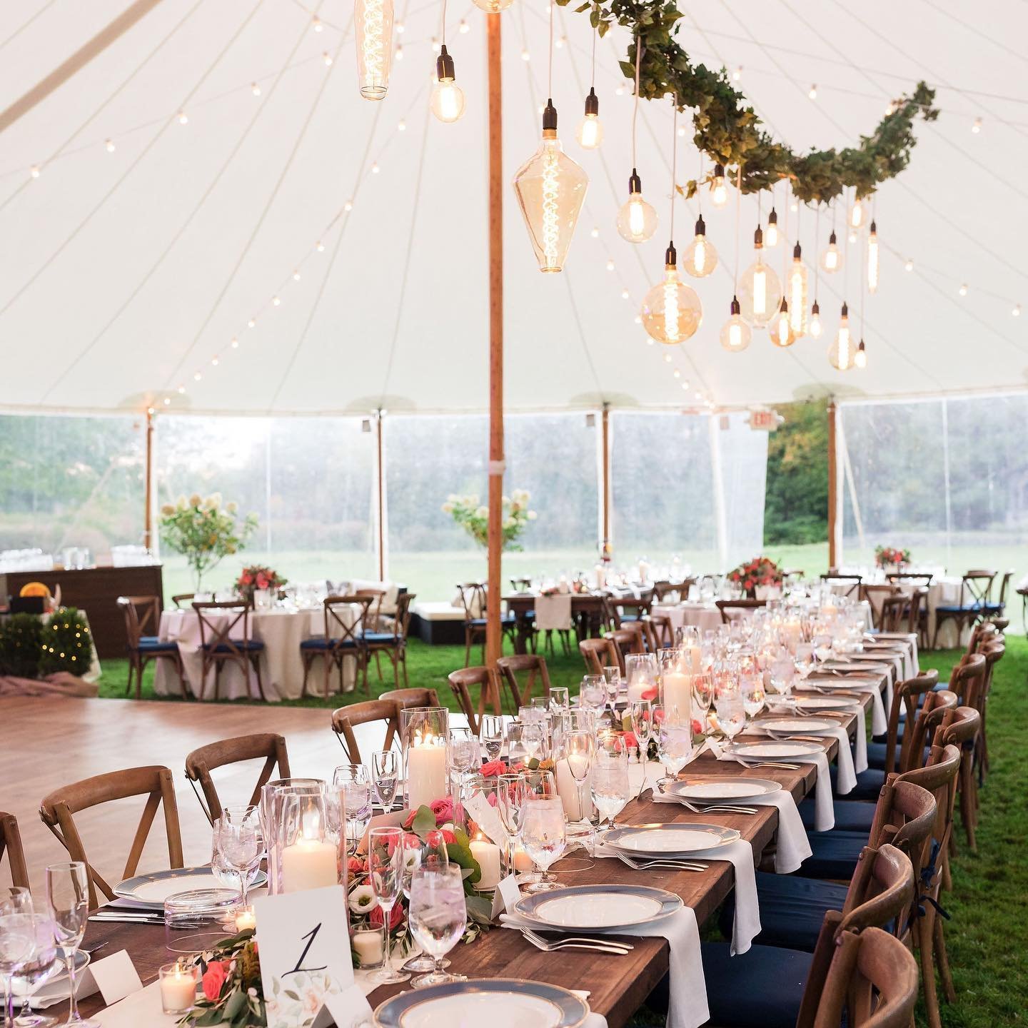 A decorated outdoor tent for a formal event with a long table set for dinner, surrounded by wooden chairs, with floral arrangements, candles, and glassware, with additional tables and floral decorations inside the tent.