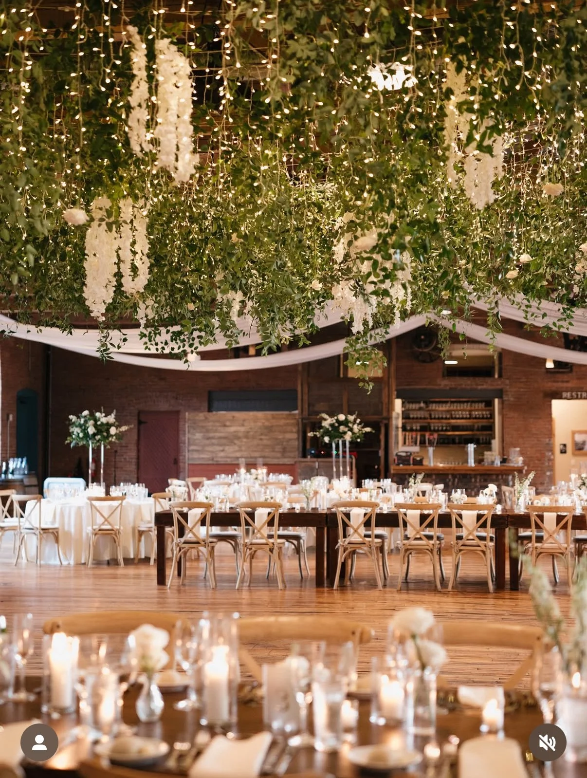 Elegant event venue decorated with hanging greenery, white flowers, and string lights, featuring round and rectangular tables with floral centerpieces, candles, and wooden chairs with X-shaped backs.