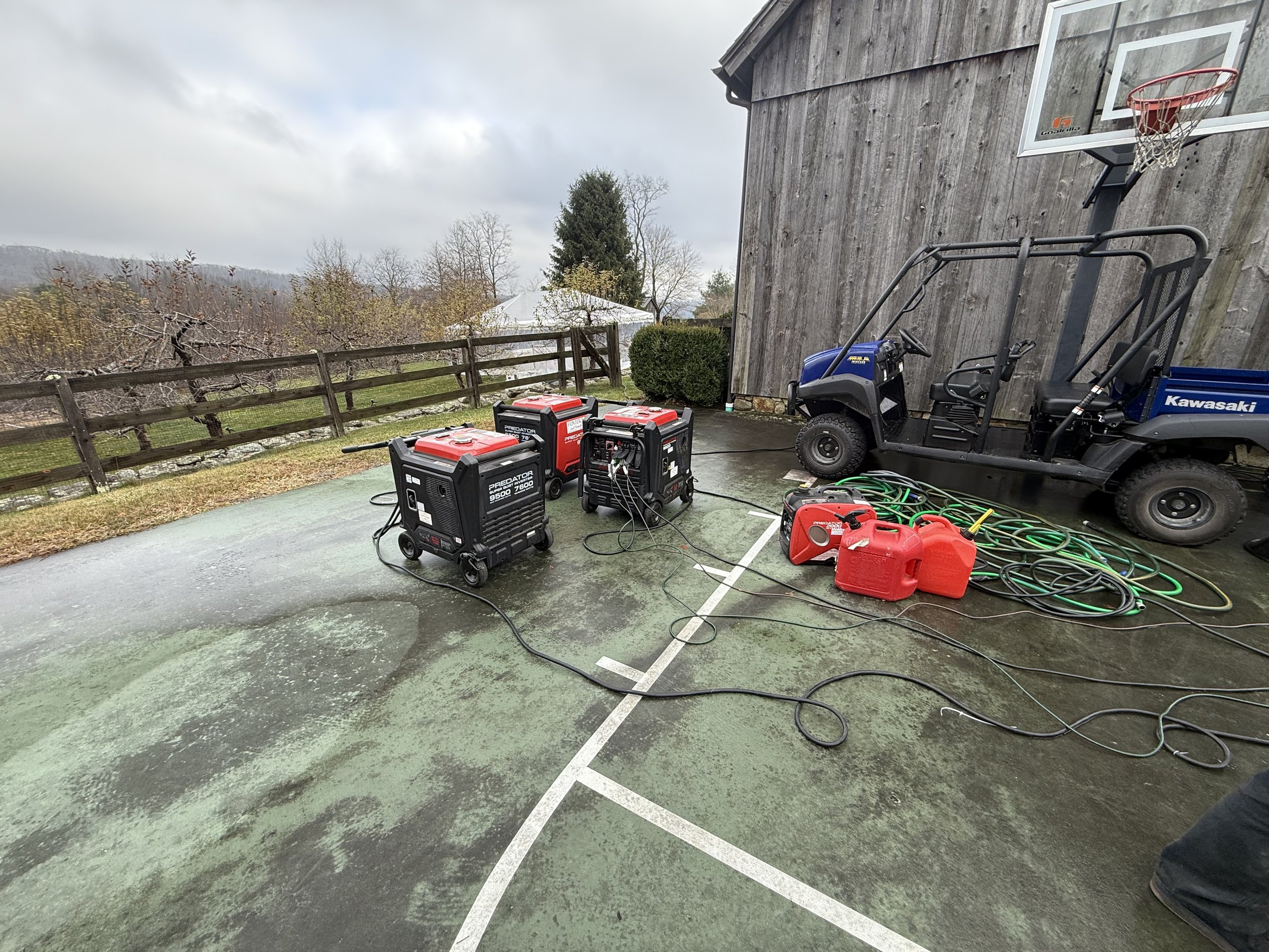 Outdoor scene with multiple welding machines, a Kawasaki utility vehicle, and red fuel containers on a wet basketball court next to a wooden building, with a rural background including trees and a cloudy sky.