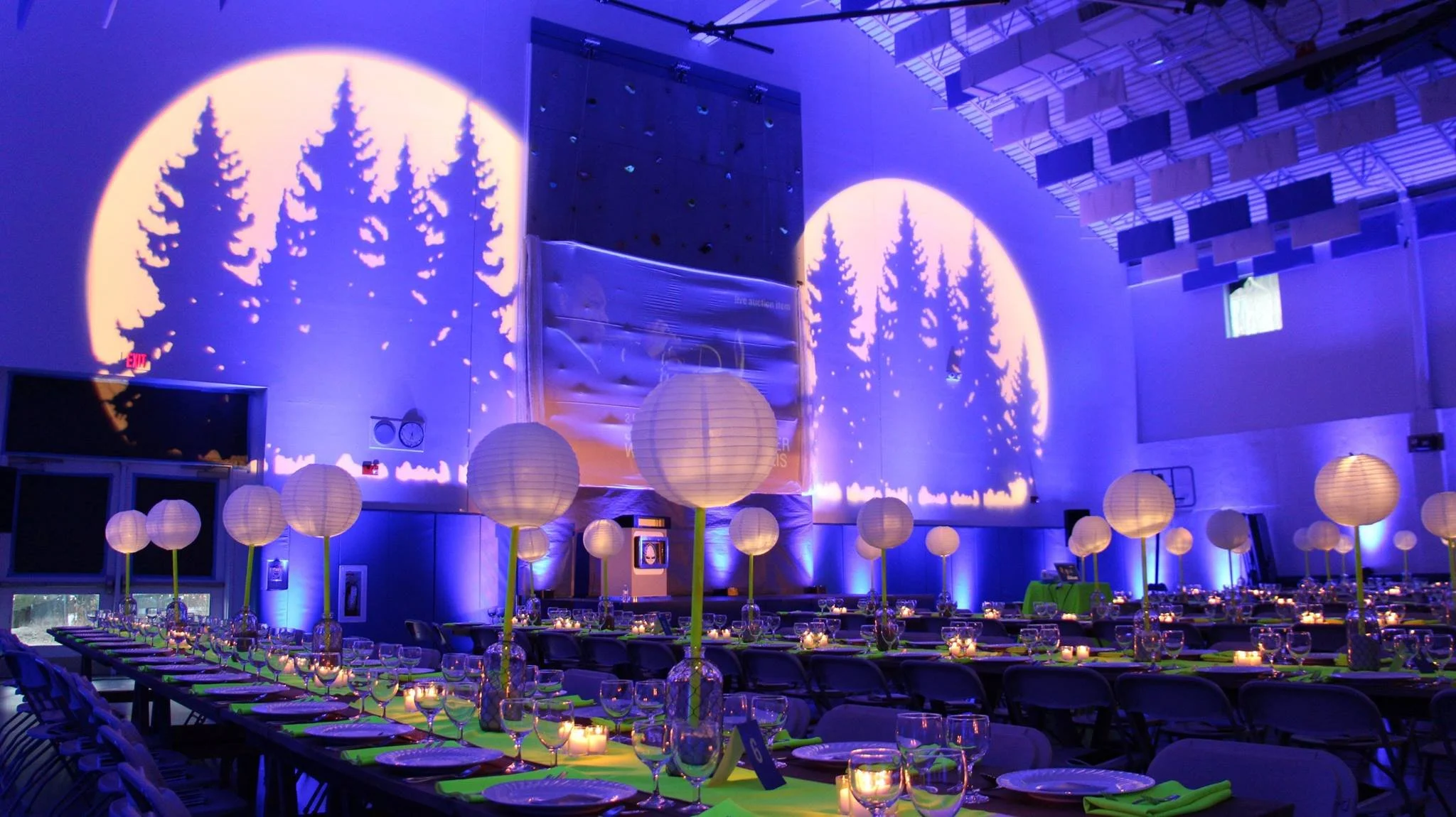 Event hall decorated with round paper lanterns, tables set for dinner with candles, blue uplighting on walls, and tree shadow projections on the walls.