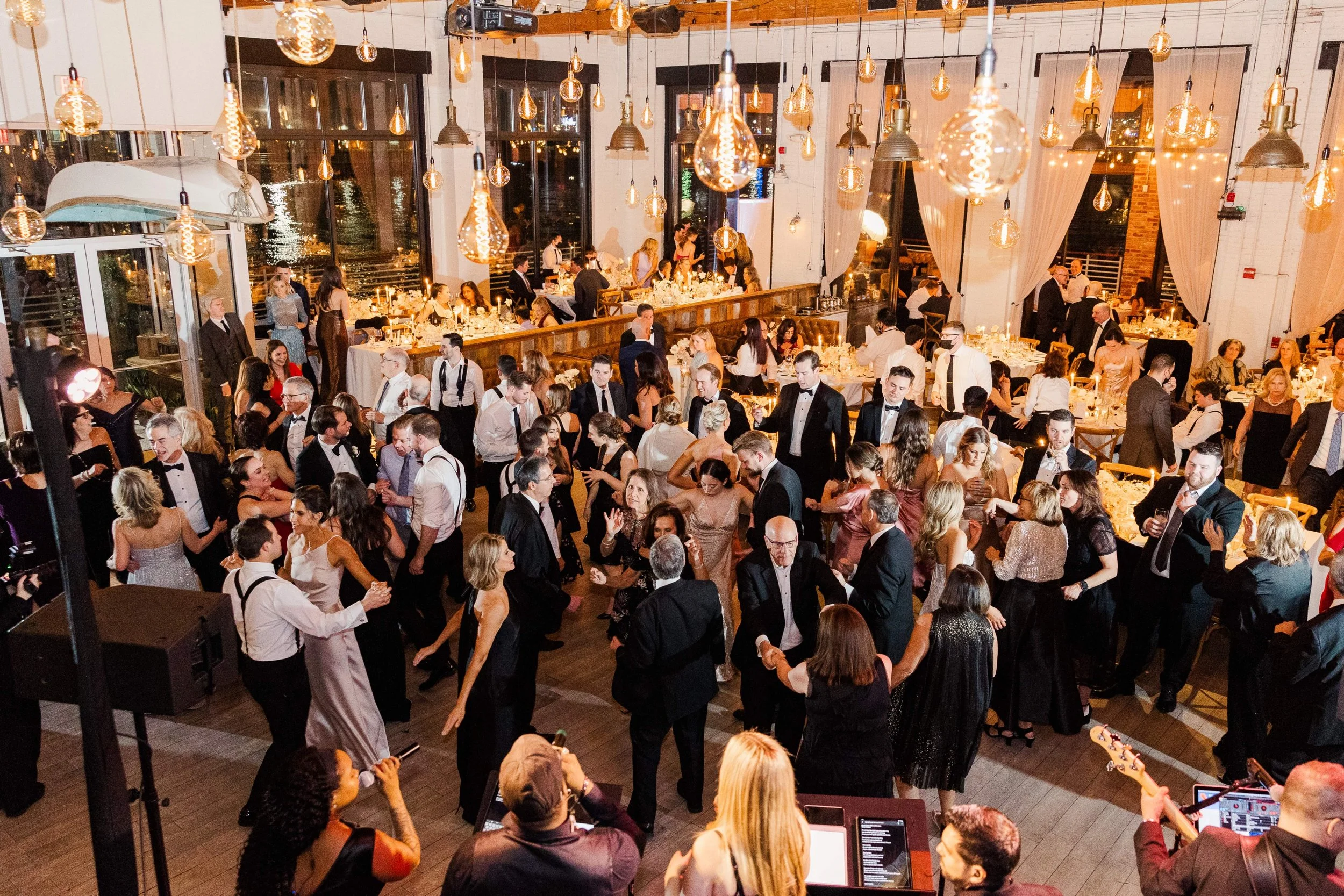 Guests dancing and socializing at a formal event or wedding reception in a decorated venue with hanging lights and tables.