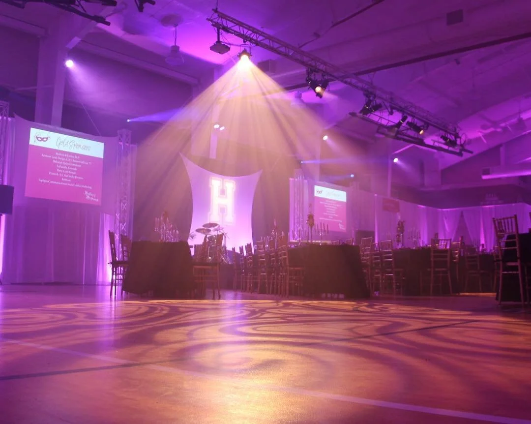 Empty banquet hall decorated with purple lighting, round tables with chairs, large white letter 'H' on stage, screens on walls displaying text, and a drum set on stage.