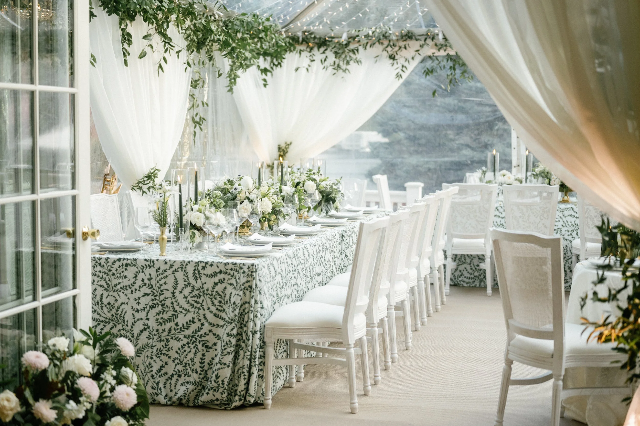 Elegant outdoor dining setup with a long table draped in a patterned cloth and decorated with white flowers, green foliage, and candles, under sheer white curtains.