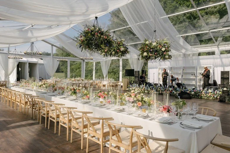 Elegant indoor wedding reception setup with long head table decorated with floral arrangements, surrounded by modern wooden chairs. There are hanging flower chandeliers, white drapery, and a live band performing in the background, with large windows showcasing greenery outside.