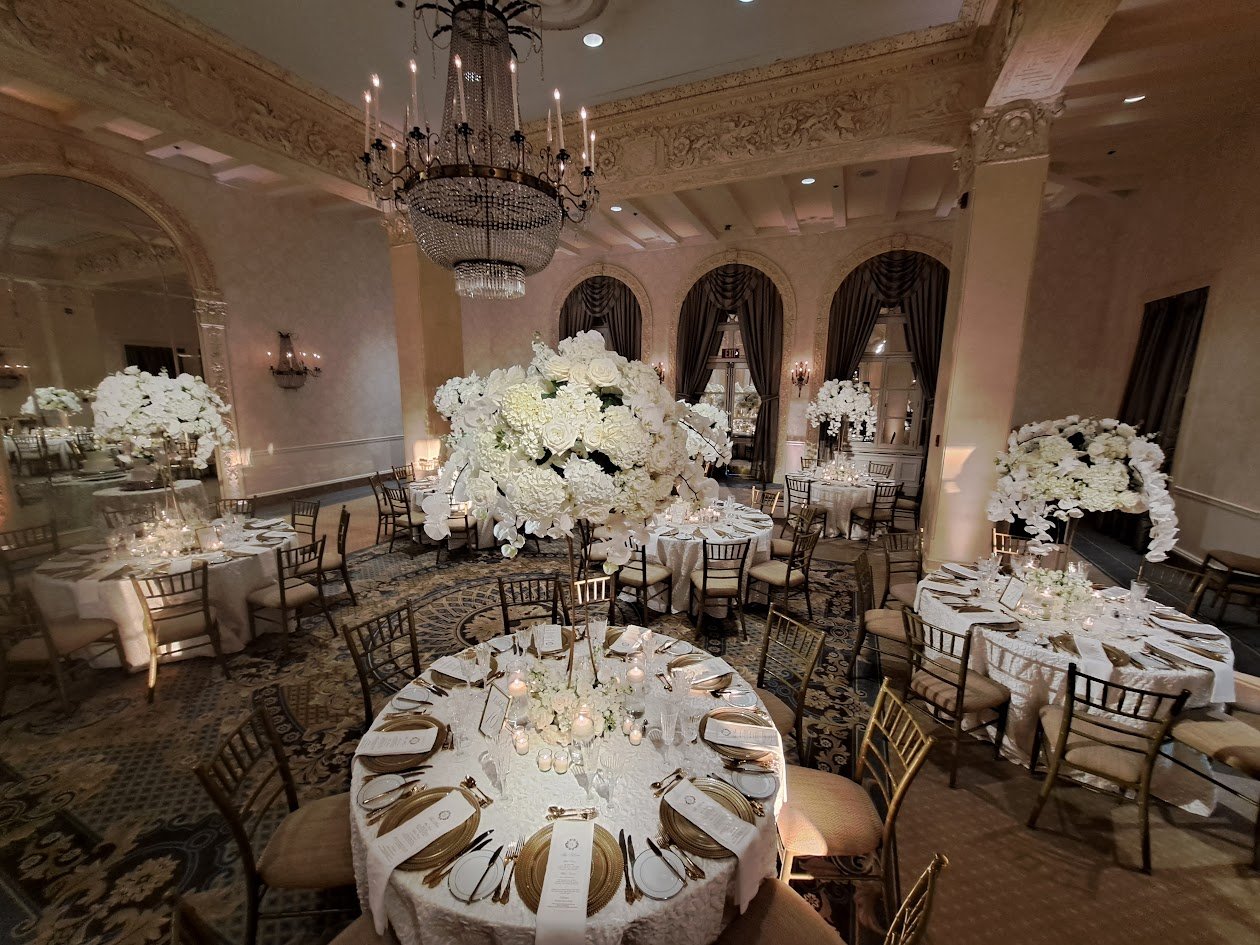 Elegant banquet hall decorated for a wedding or special event with round tables adorned with white tablecloths, floral centerpieces of white roses and hydrangeas, gold charger plates, and gold-colored cutlery. Multiple floral arrangements and tall candles are on the tables, with a grand chandelier hanging from the ceiling and large, dark curtains framing windows in the background.