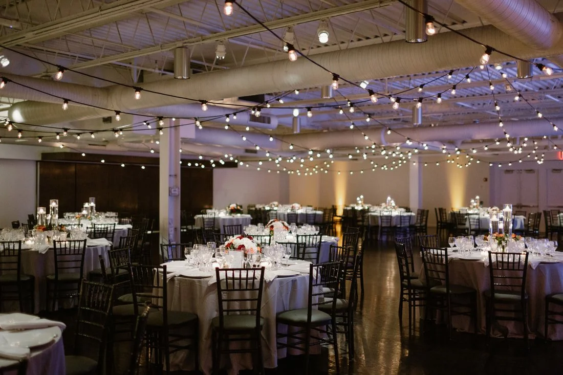 Decorated banquet hall with round tables, floral centerpieces, glassware, and string lights hanging from the ceiling.
