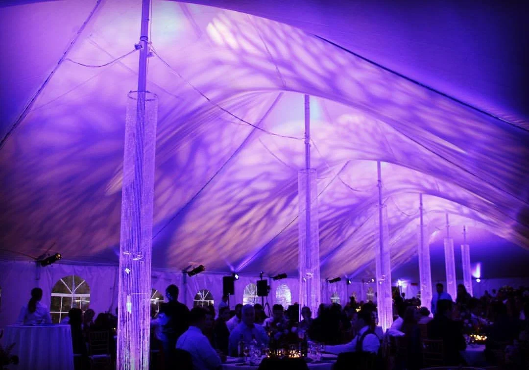 Inside a large event tent with purple lighting, decorated with hanging crystal chandeliers, where guests are seated at tables for a celebration or reception.