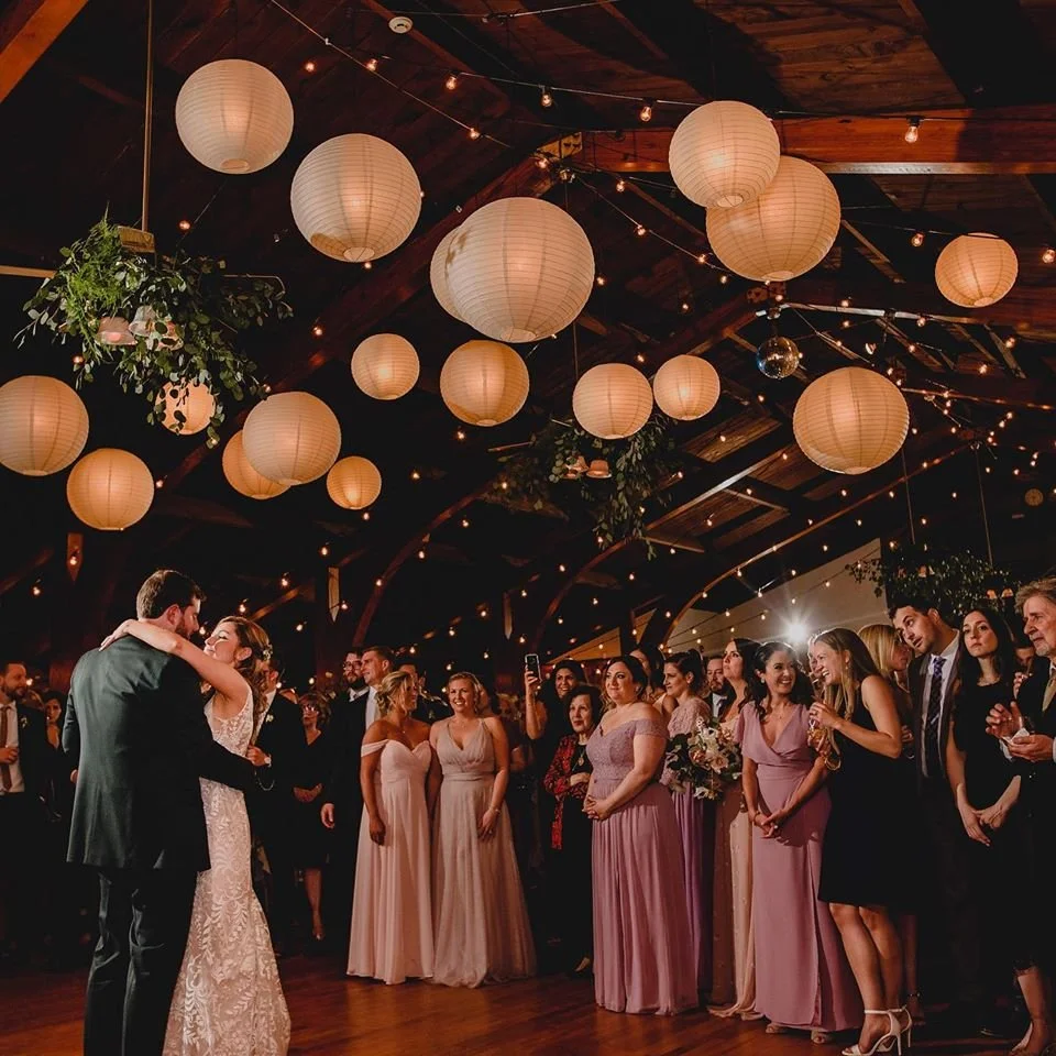 Bride and groom dancing at their wedding reception with guests watching in a decorated venue with paper lanterns and string lights.