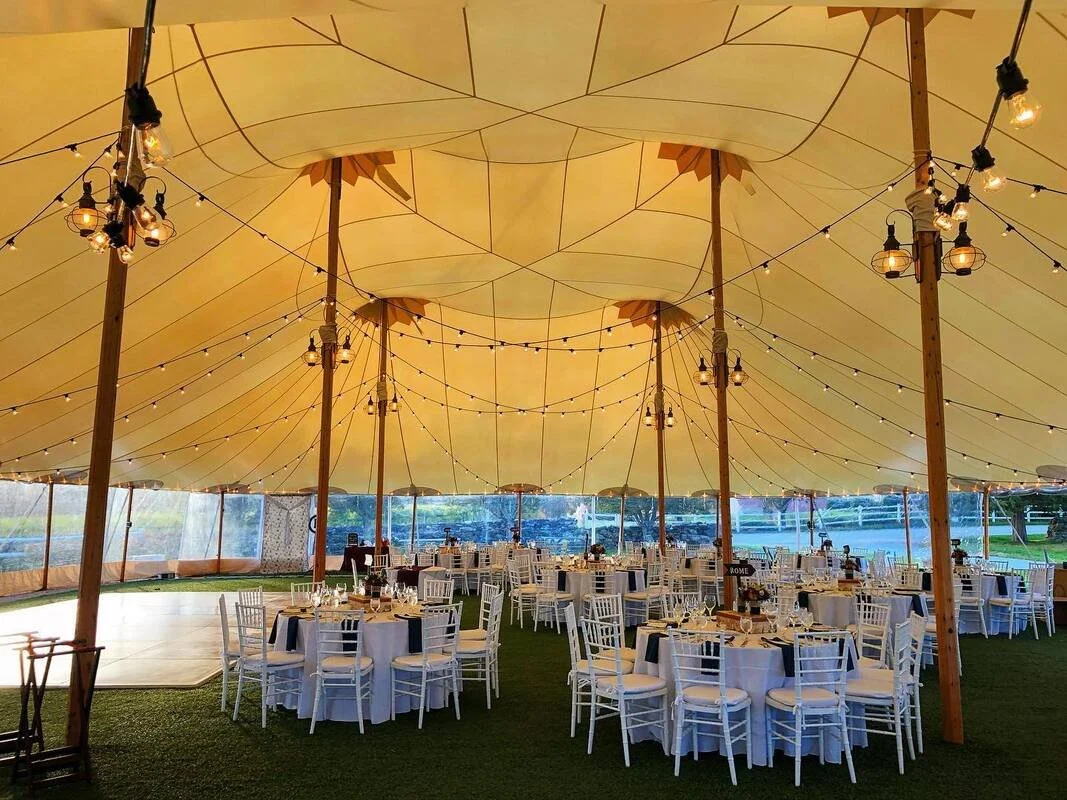 A large event tent decorated with string lights, with round tables covered in navy blue and white tablecloths, set for a banquet or celebration, with white chairs around each table, on a grassy area.