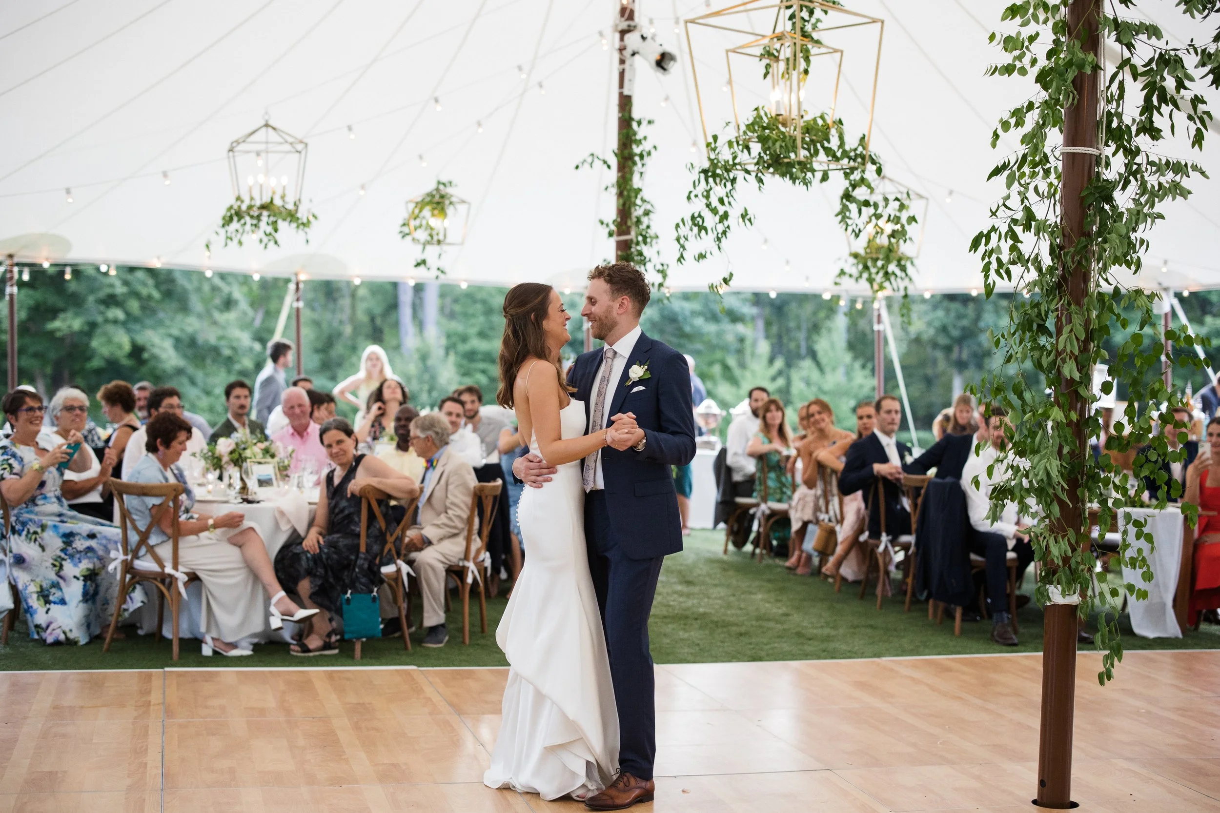 A decorated event space with hanging white floral arrangements and string lights from the ceiling. The room has round and rectangular tables with white tablecloths and floral centerpieces, set for a celebration or wedding, with wooden chairs and a brick wall in the background.