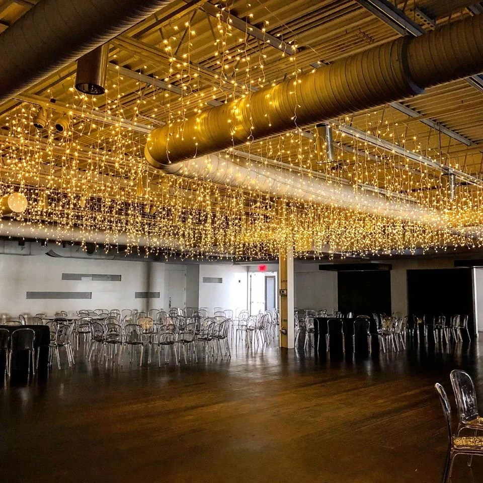 An indoor event space decorated with string lights hanging from the ceiling, with clear plastic chairs arranged around tables, and a wooden floor.