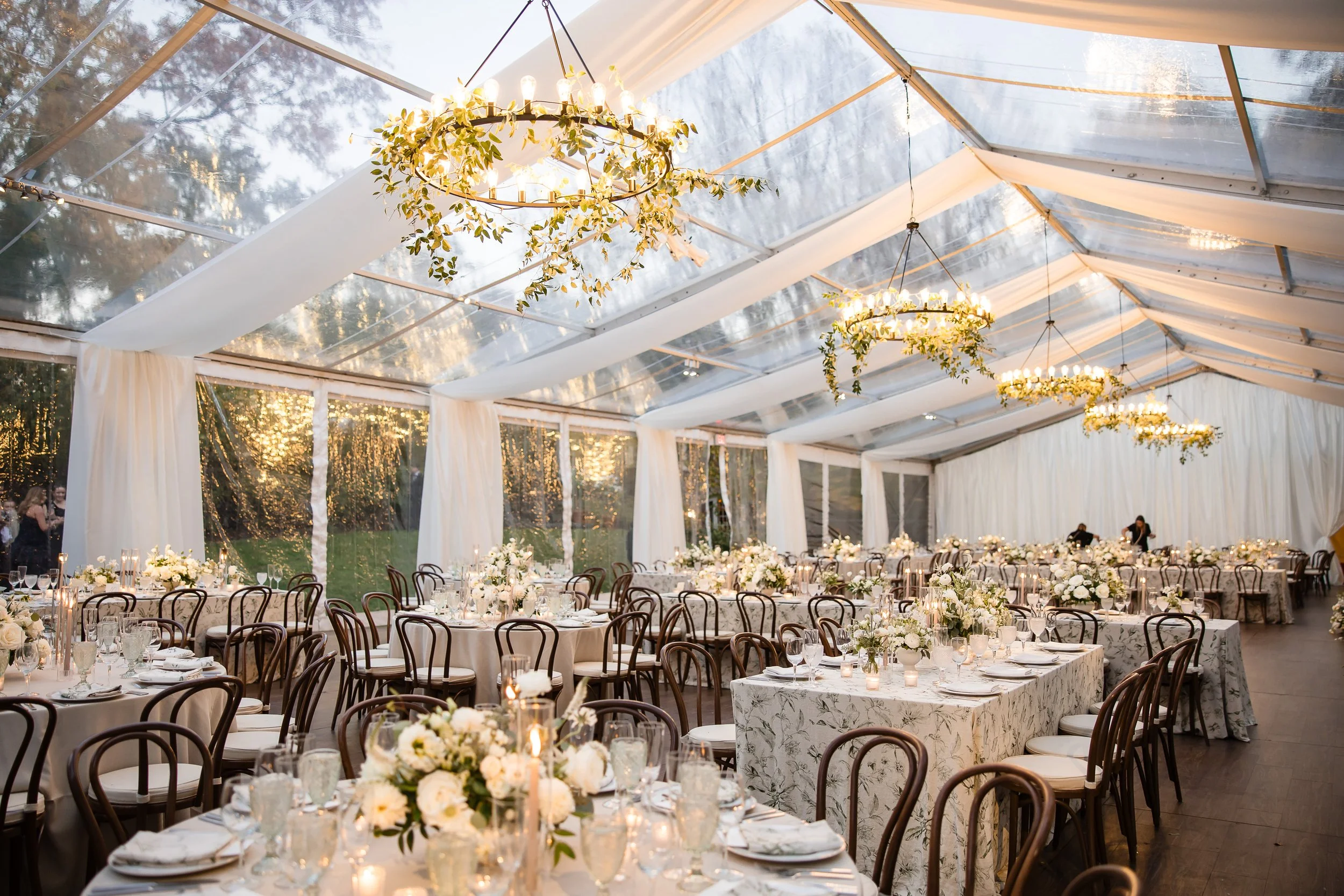 Elegant indoor wedding reception with round tables, floral centerpieces, and hanging chandeliers decorated with greenery in a clear tent.