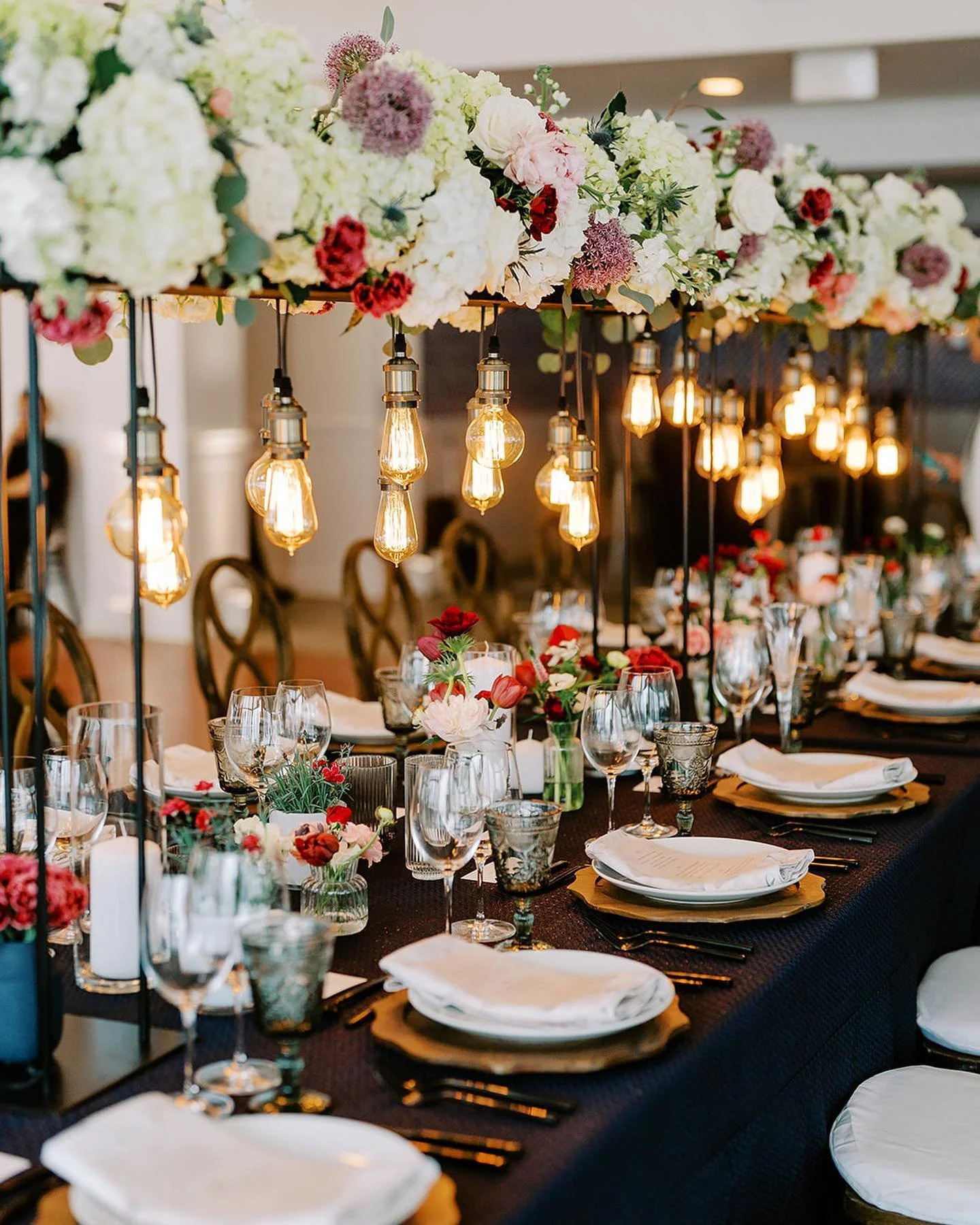 Elegant banquet table decorated with floral centerpieces, hanging Edison-style light bulbs, and set with glassware, plates, forks, and napkins.