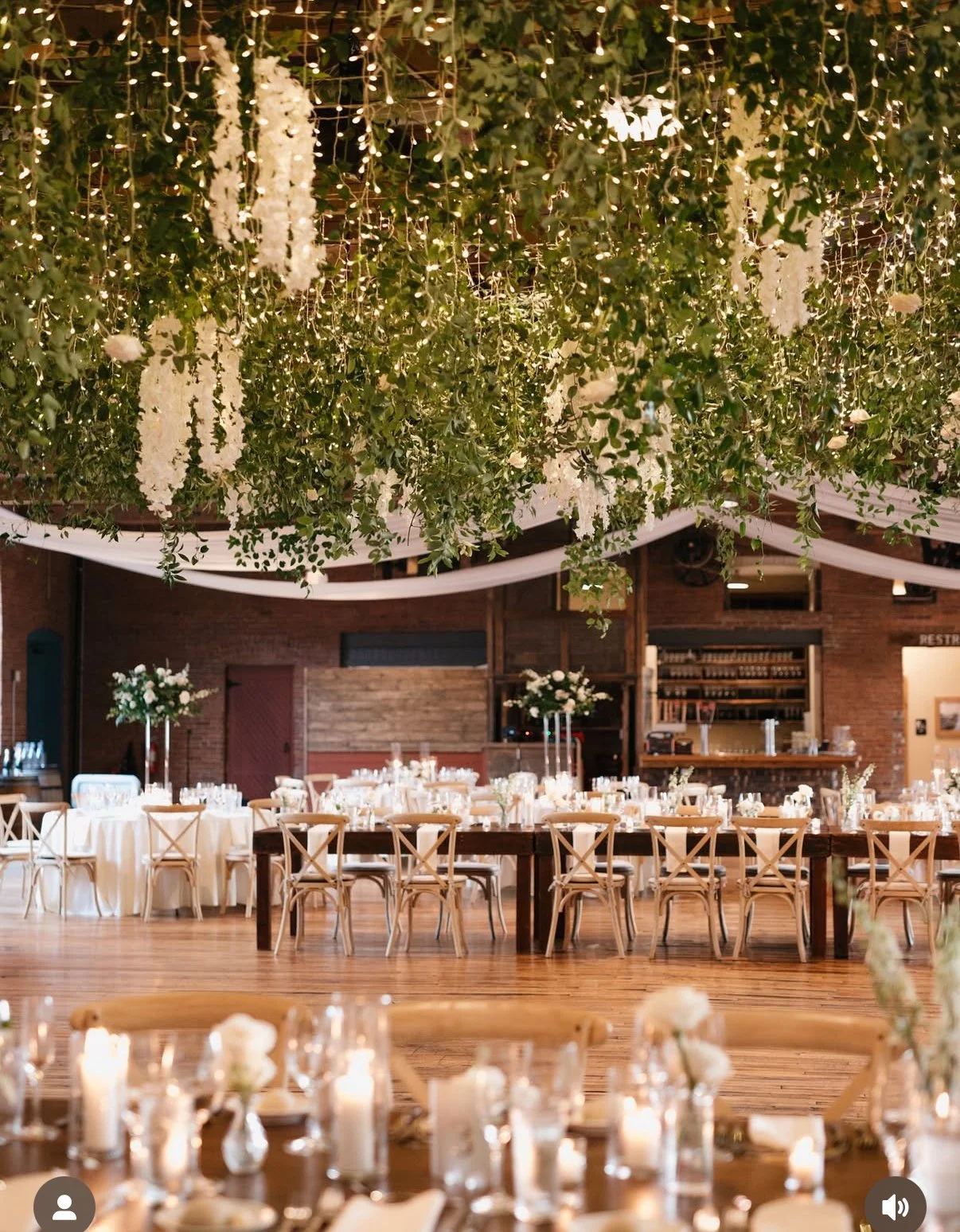 A decorated event space with hanging white floral arrangements and string lights from the ceiling. The room has round and rectangular tables with white tablecloths and floral centerpieces, set for a celebration or wedding, with wooden chairs and a brick wall in the background.