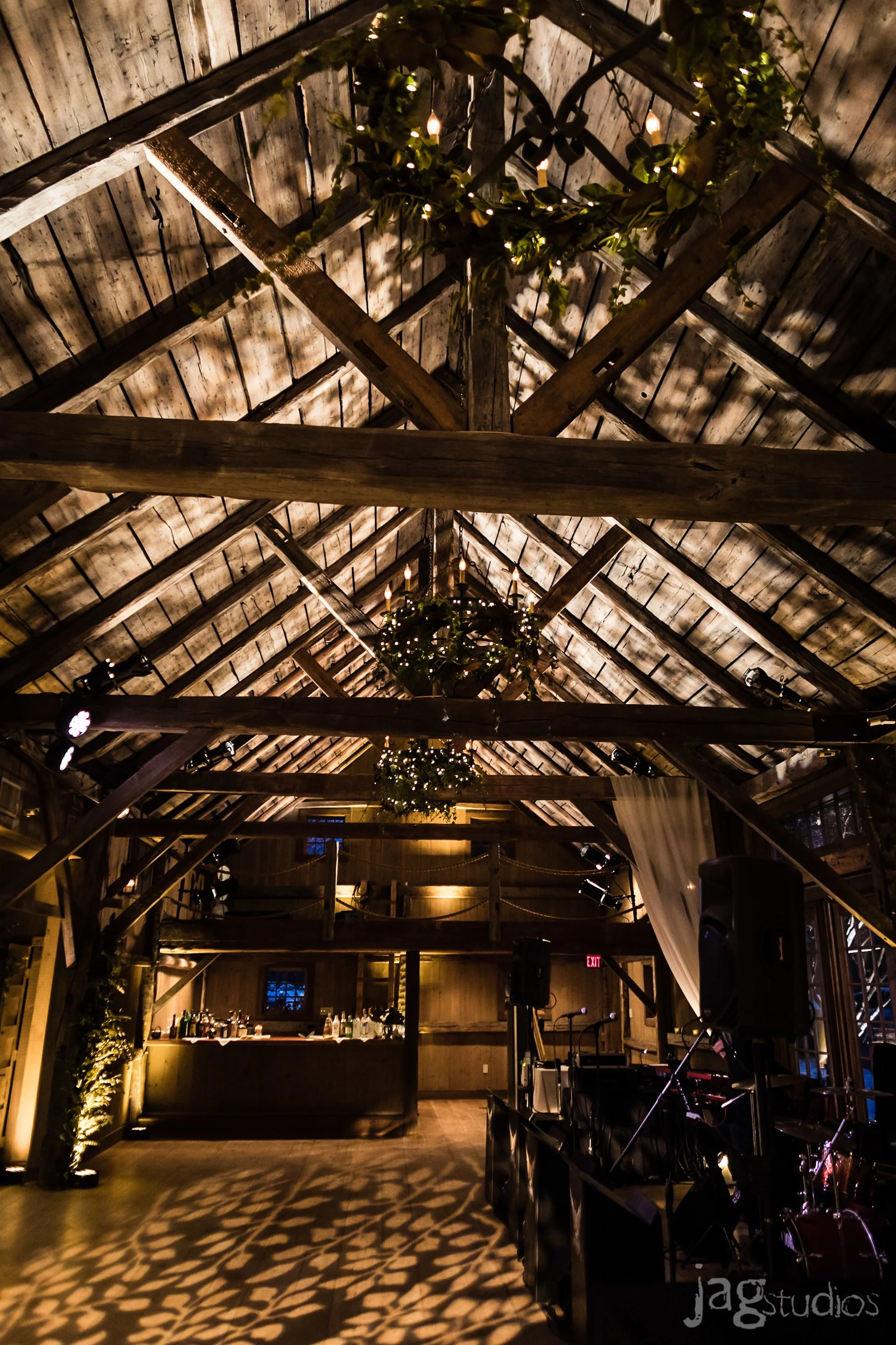 Interior of a rustic wedding barn with wooden beams, chandeliers decorated with greenery and lights, a dance floor with patterned shadows, a bar area, and a music setup.