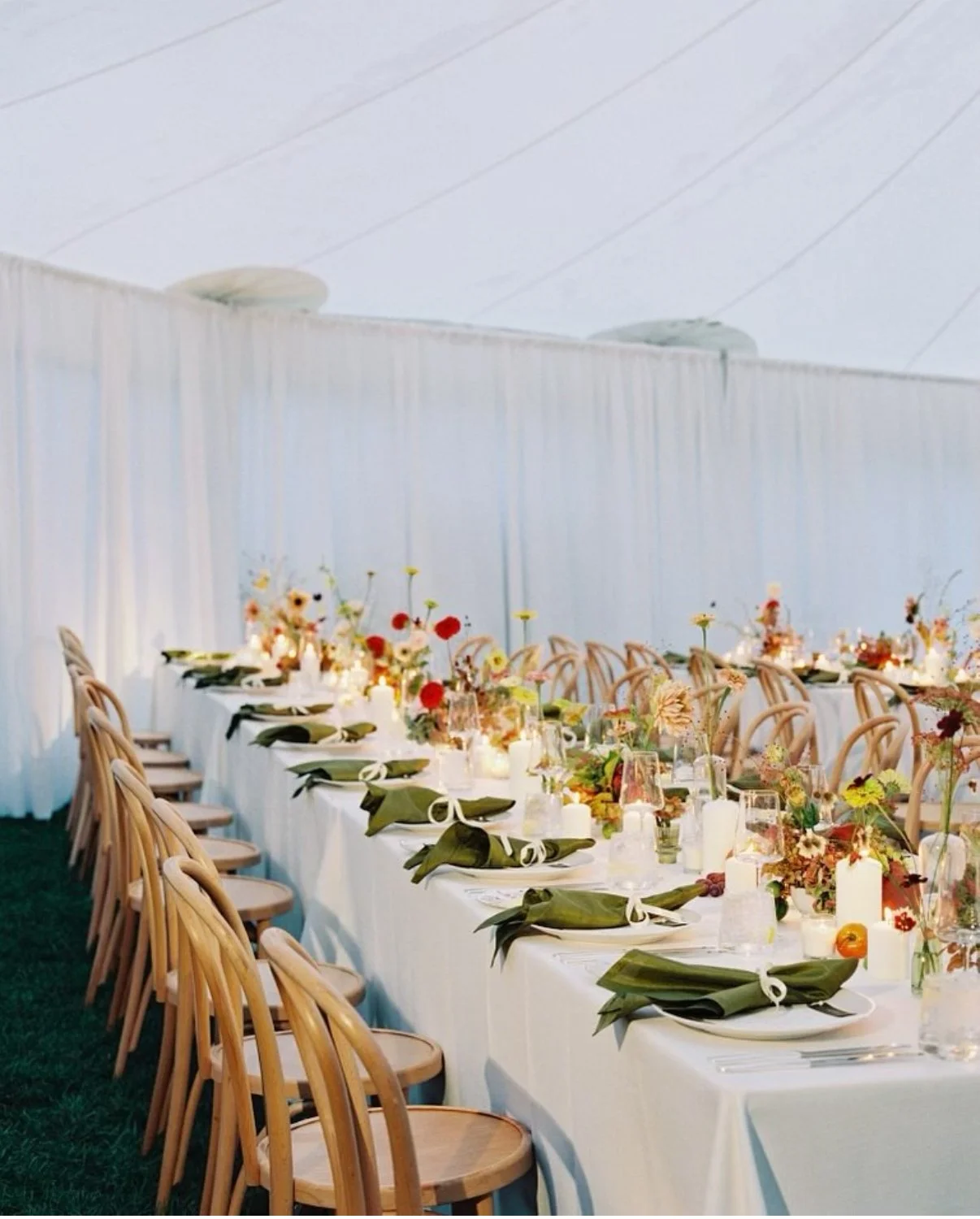Long outdoor dining table with white tablecloth, decorated with floral centerpieces, candles, and place settings, set under a white canopy.