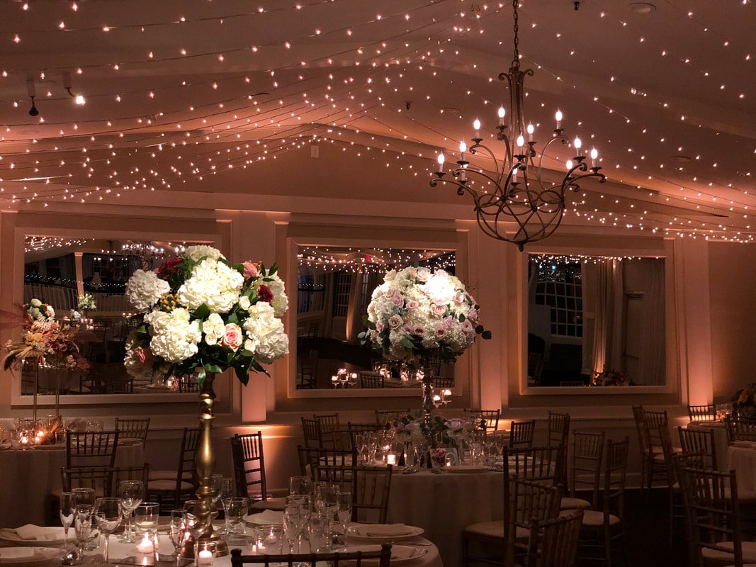 A decorated banquet hall with floral centerpieces, string lights on the ceiling, and a chandelier, set for an elegant event.