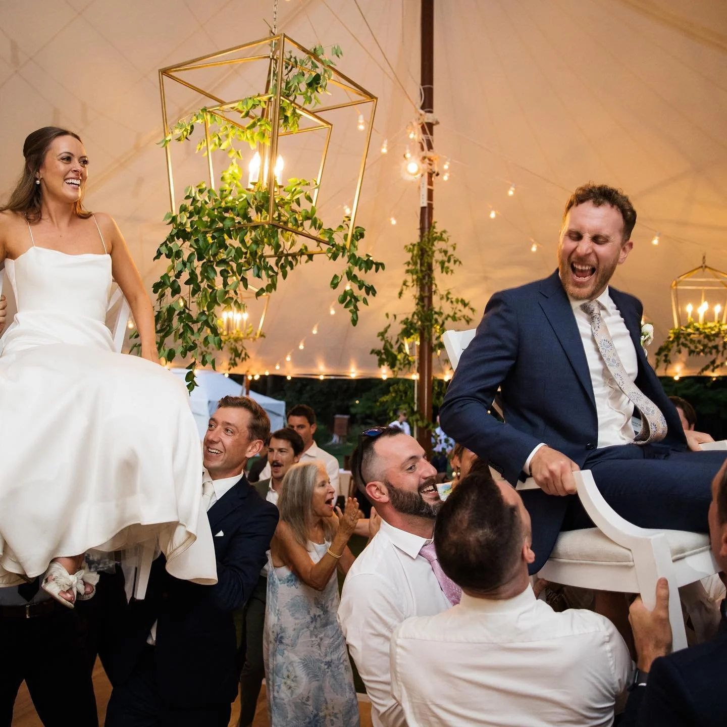 People at a wedding celebration lifting the bride and groom, smiling and laughing under a decorated tent with hanging lights and greenery.