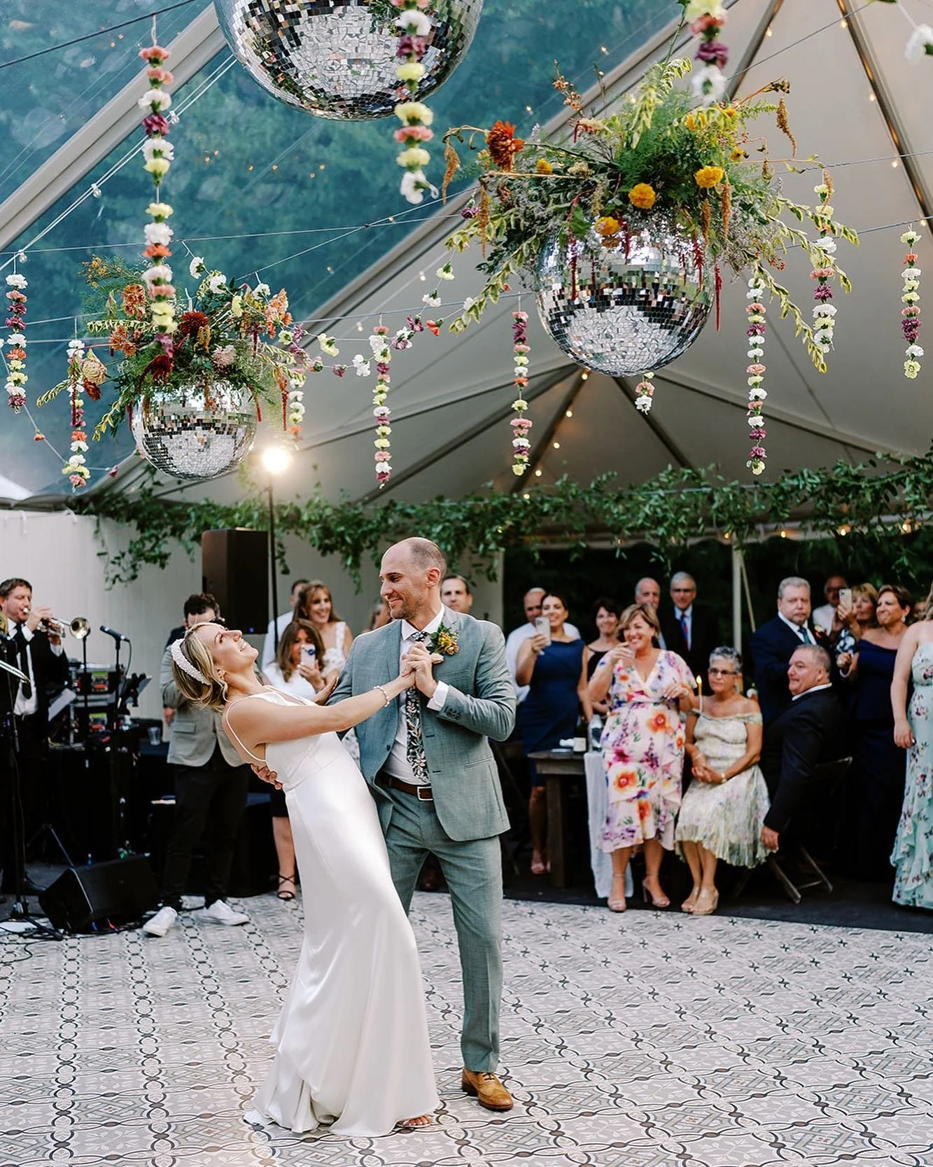 A bride and groom dancing at their wedding reception, surrounded by guests, with floral arrangements and disco balls hanging from the ceiling.